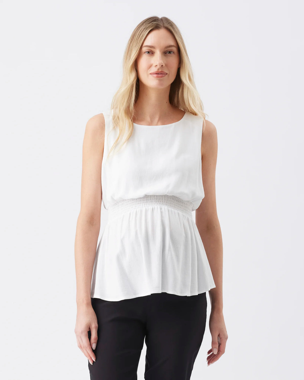 A blonde woman modeling a sleeveless white peplum top with a smocked waistband, paired with black pants, against a plain white studio background.