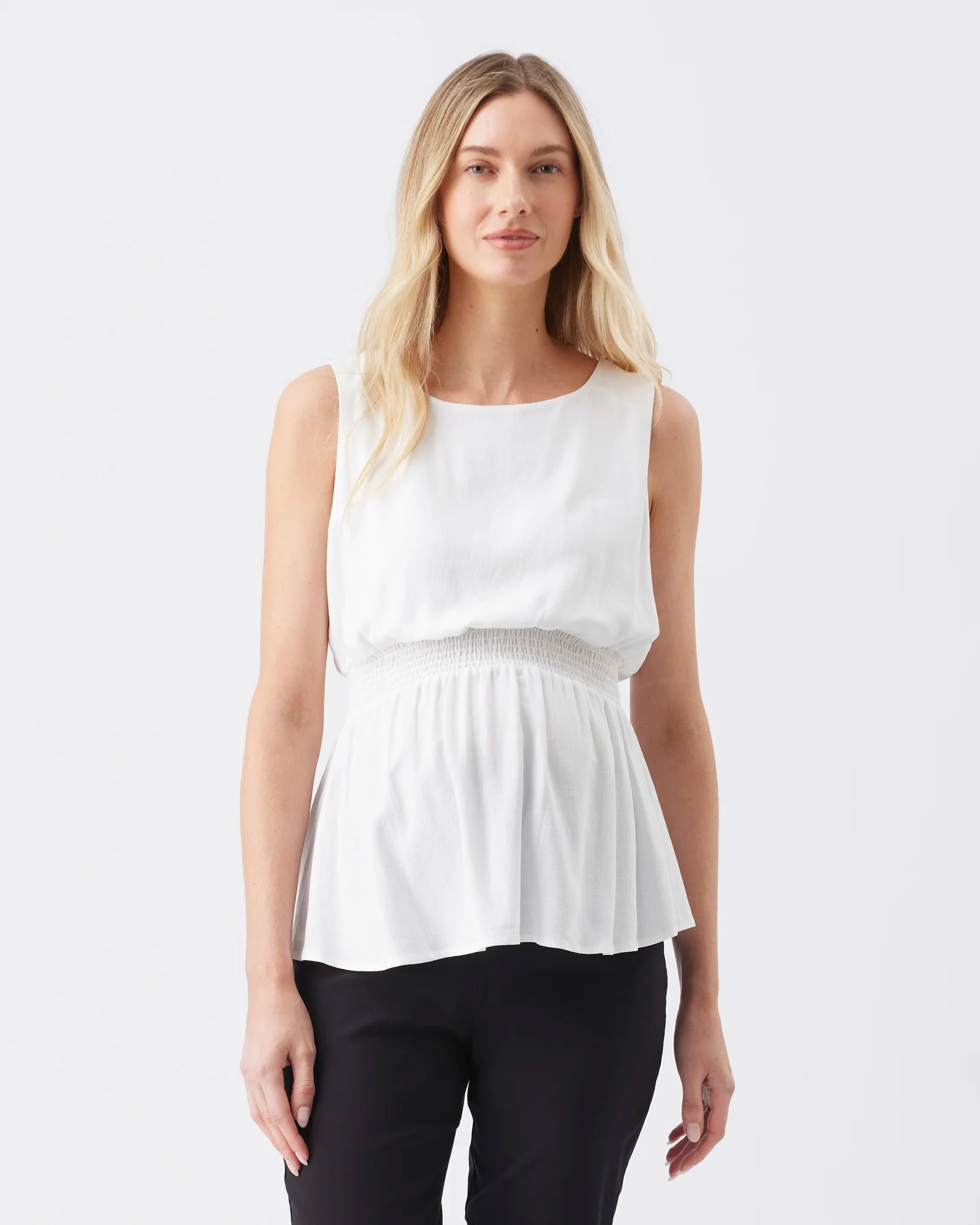 A blonde woman modeling a sleeveless white peplum top with a smocked waistband, paired with black pants, against a plain white studio background.