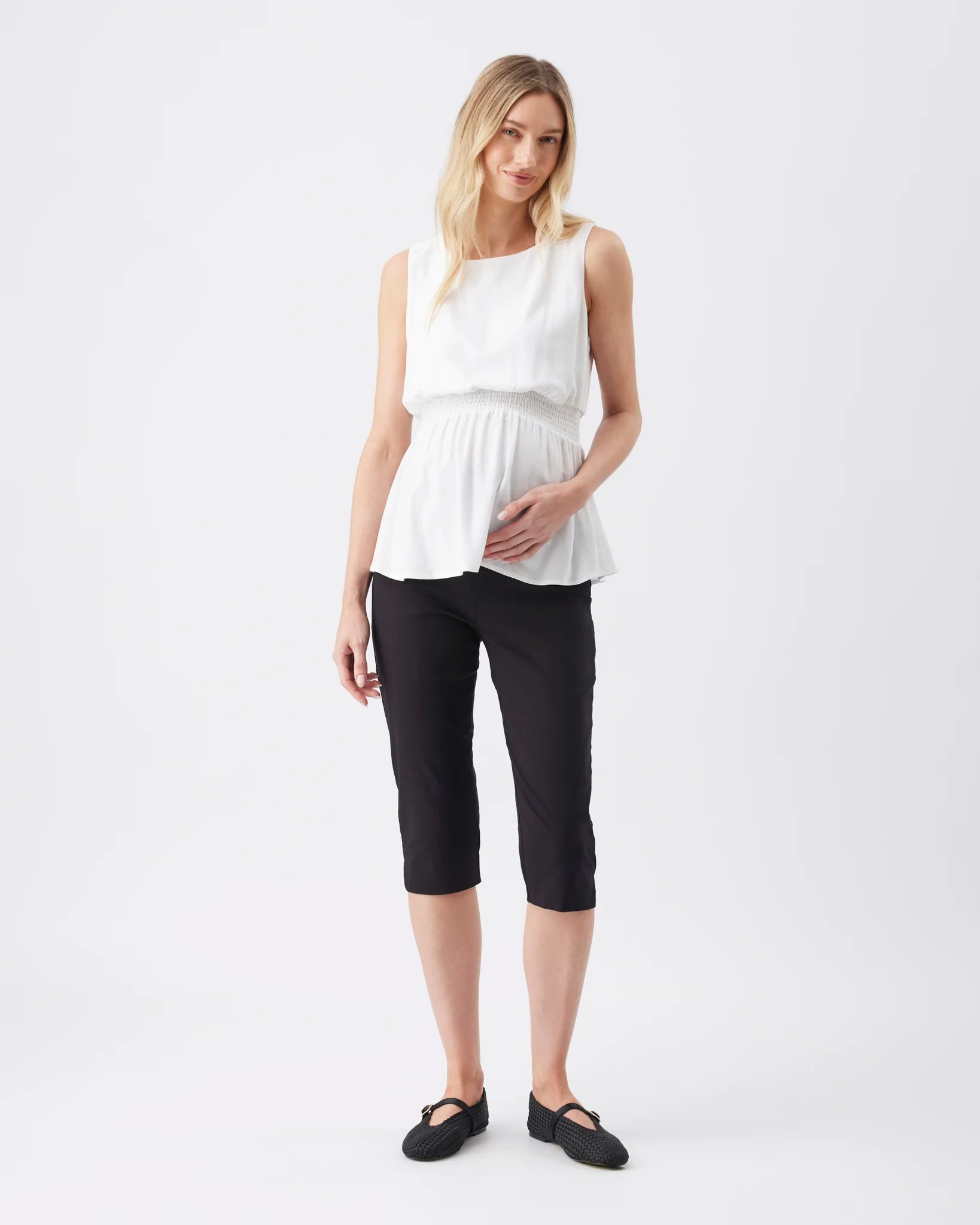 A woman modeling a white sleeveless peplum top and black cropped pants against a plain light gray studio background.