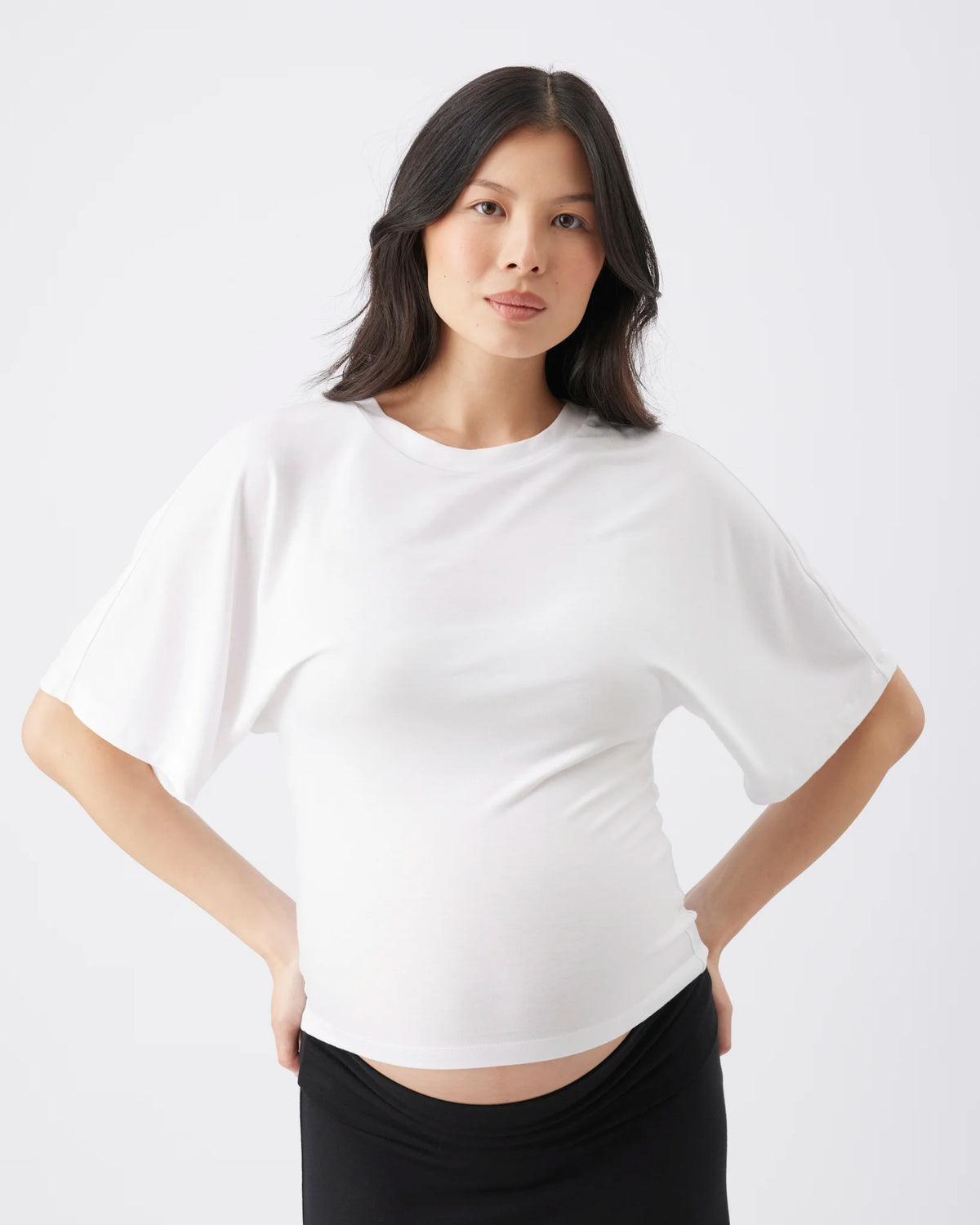 A woman modeling a white short-sleeve crewneck top with a draped, oversized silhouette, paired with black bottoms, against a plain light gray studio background.