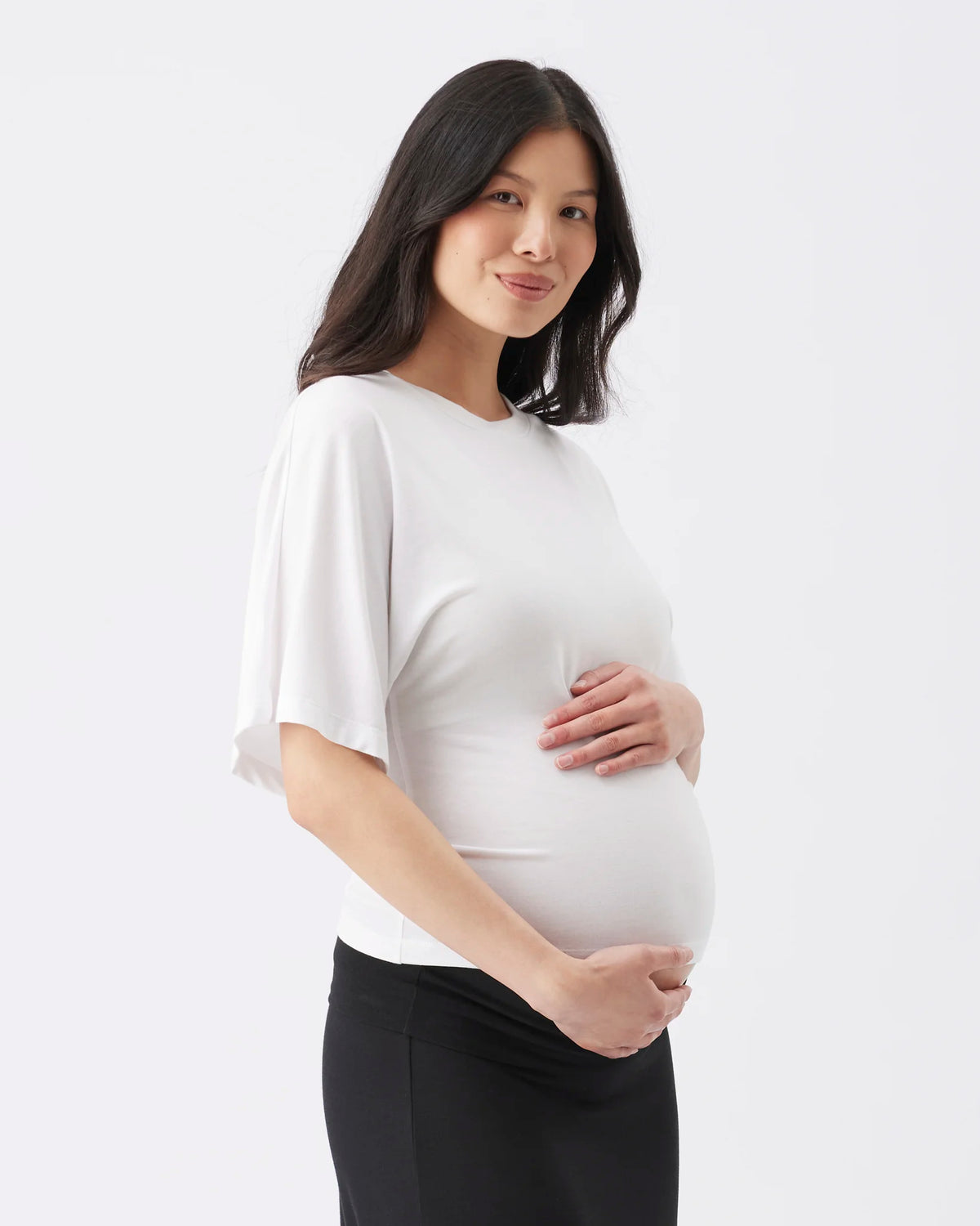 A pregnant woman with long dark hair stands in profile against a light gray studio background, wearing a loose white short-sleeve top and black pants, with her hands resting on her belly.