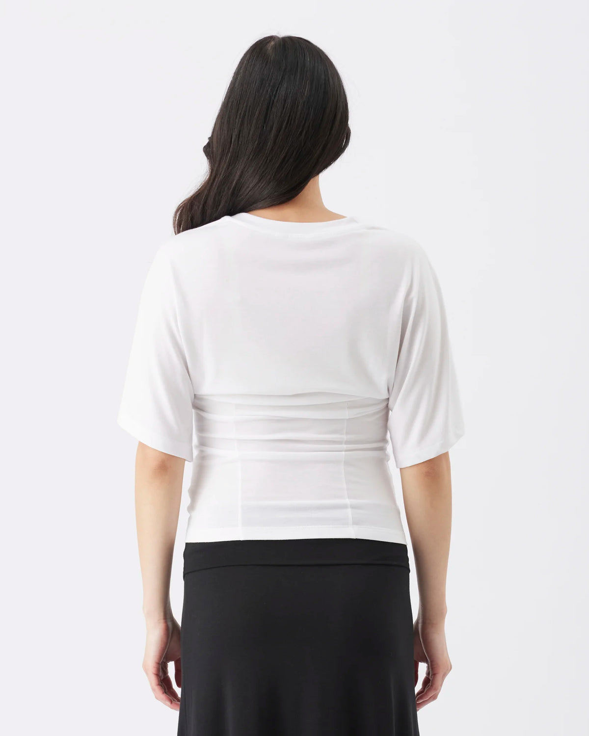 Back view of a pregnant woman against a light gray studio background, wearing a loose white short-sleeve top and black pants.