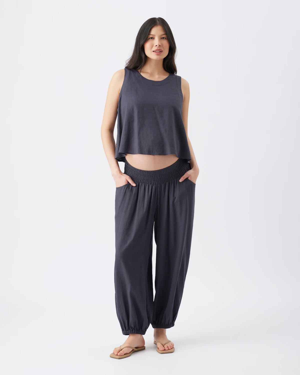 A woman modeling a dark slate gray two-piece lounge set consisting of a sleeveless cropped tank top and matching wide-leg elastic-waist pants with ankle cuffs, paired with tan sandals against a plain white studio background.