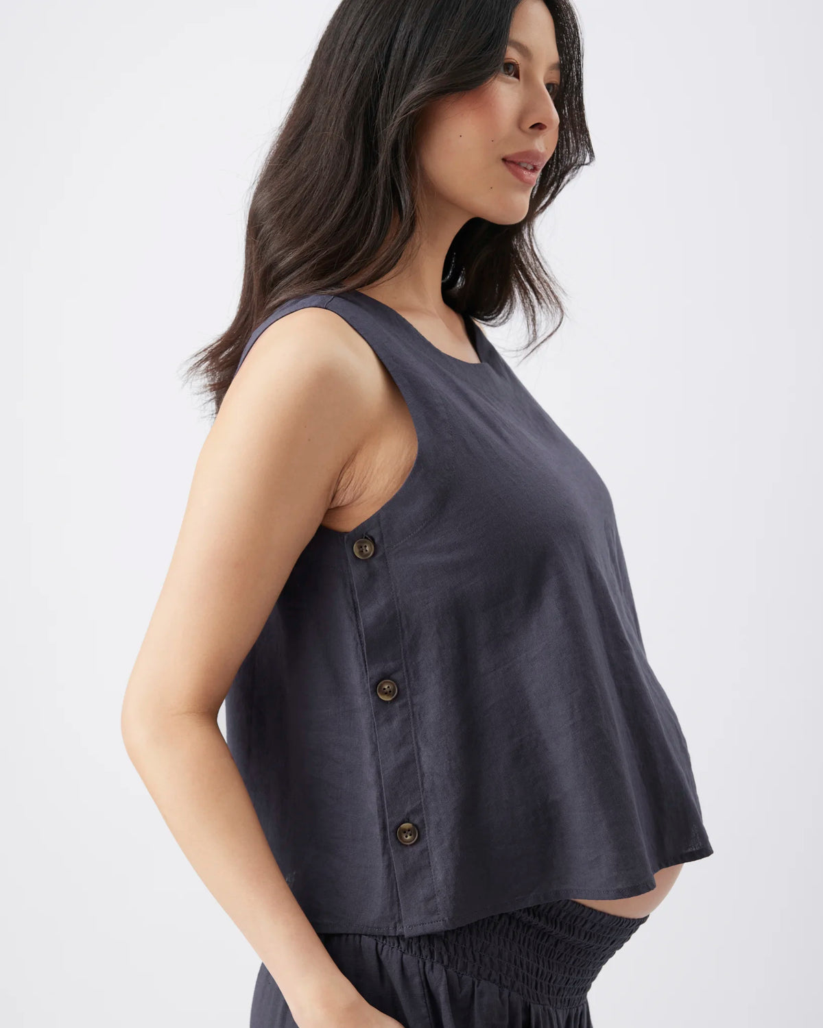 A woman wearing a sleeveless dark slate blue top with a buttoned side detail, shown in profile against a light gray studio background.
