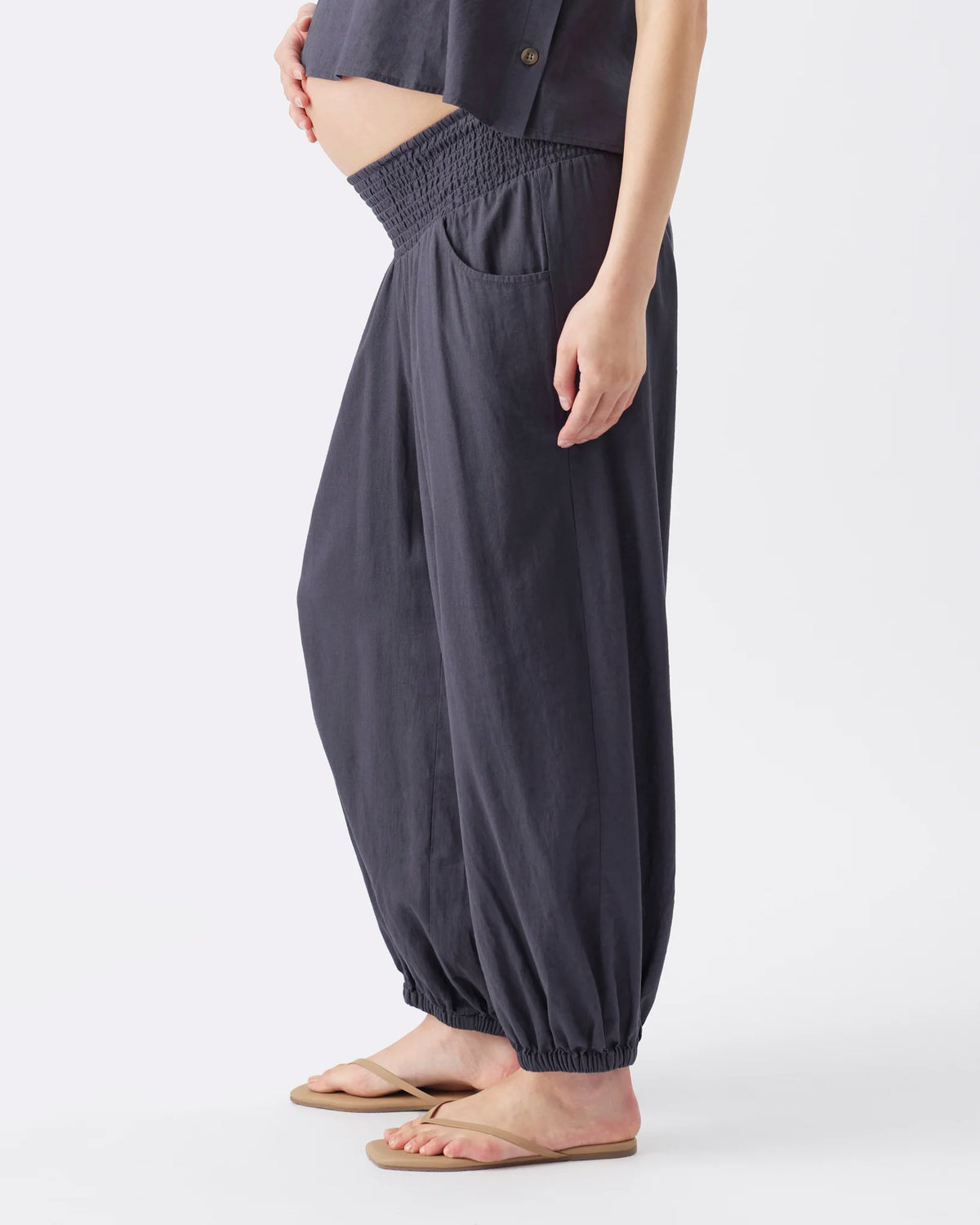 A person wearing loose, high-waisted harem-style pants in a dark charcoal gray color, paired with a matching dark top and beige flip-flop sandals, shown against a plain light gray studio background.