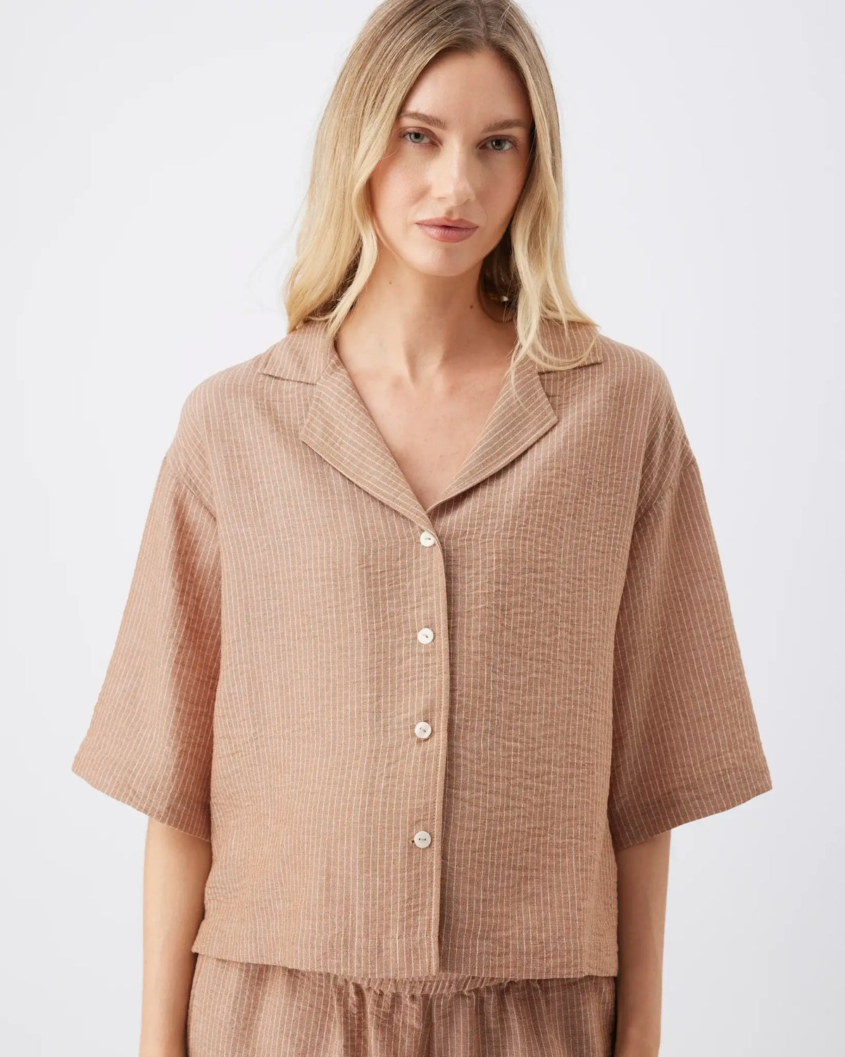 A woman wearing a short-sleeve button-front top in a textured taupe-brown fabric, photographed against a clean light gray studio background.