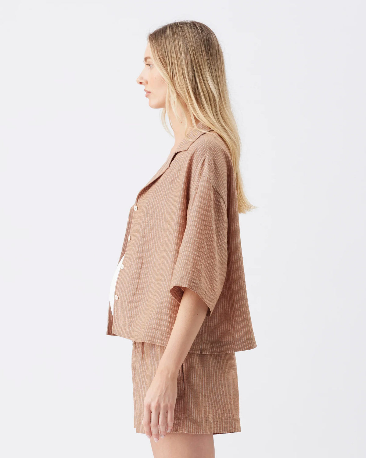 A woman in side profile wearing a matching beige textured short-sleeve button-front jacket and shorts set against a plain light gray studio background.