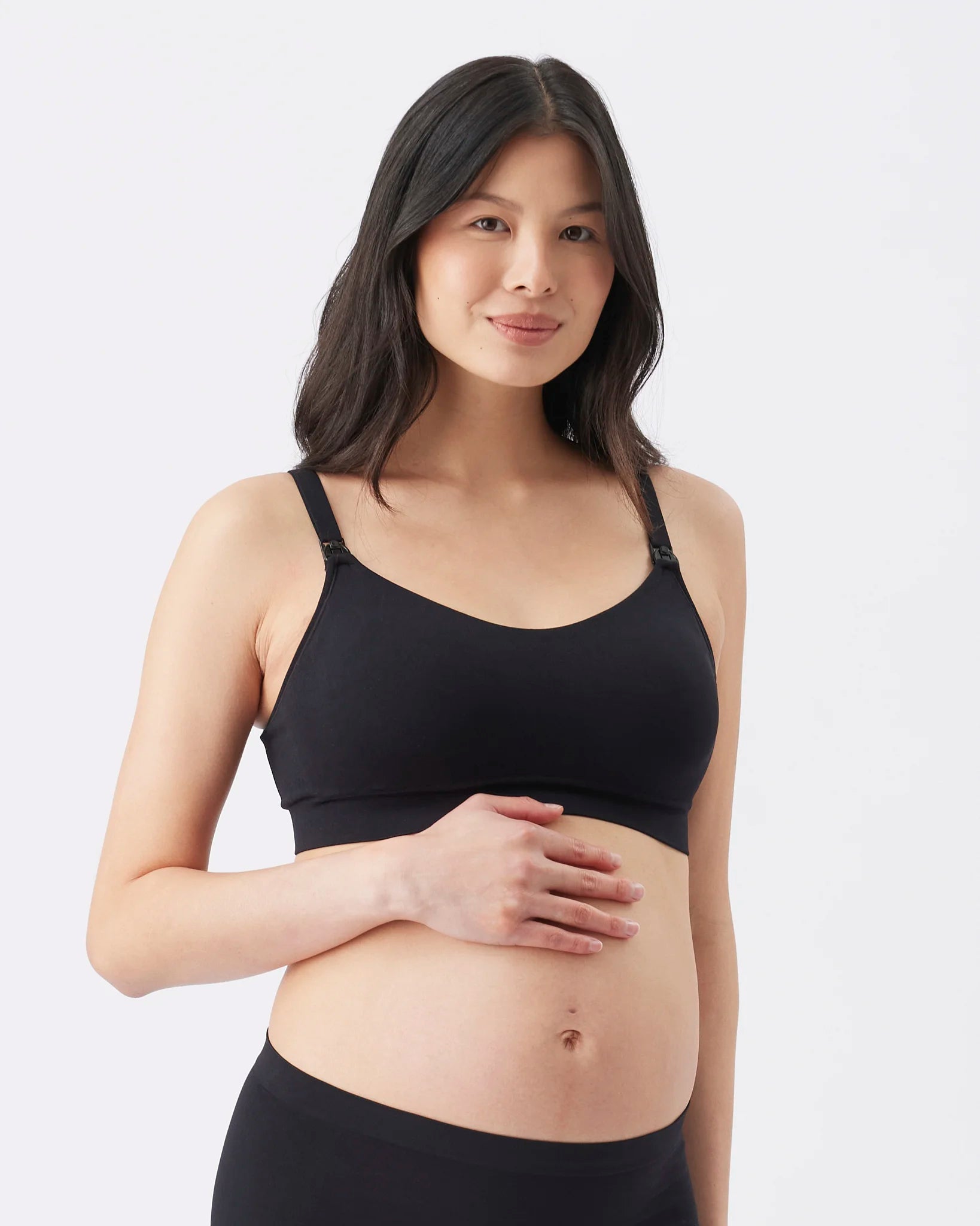 A pregnant woman wearing a black maternity nursing bra and matching black underwear, standing against a light gray studio background.