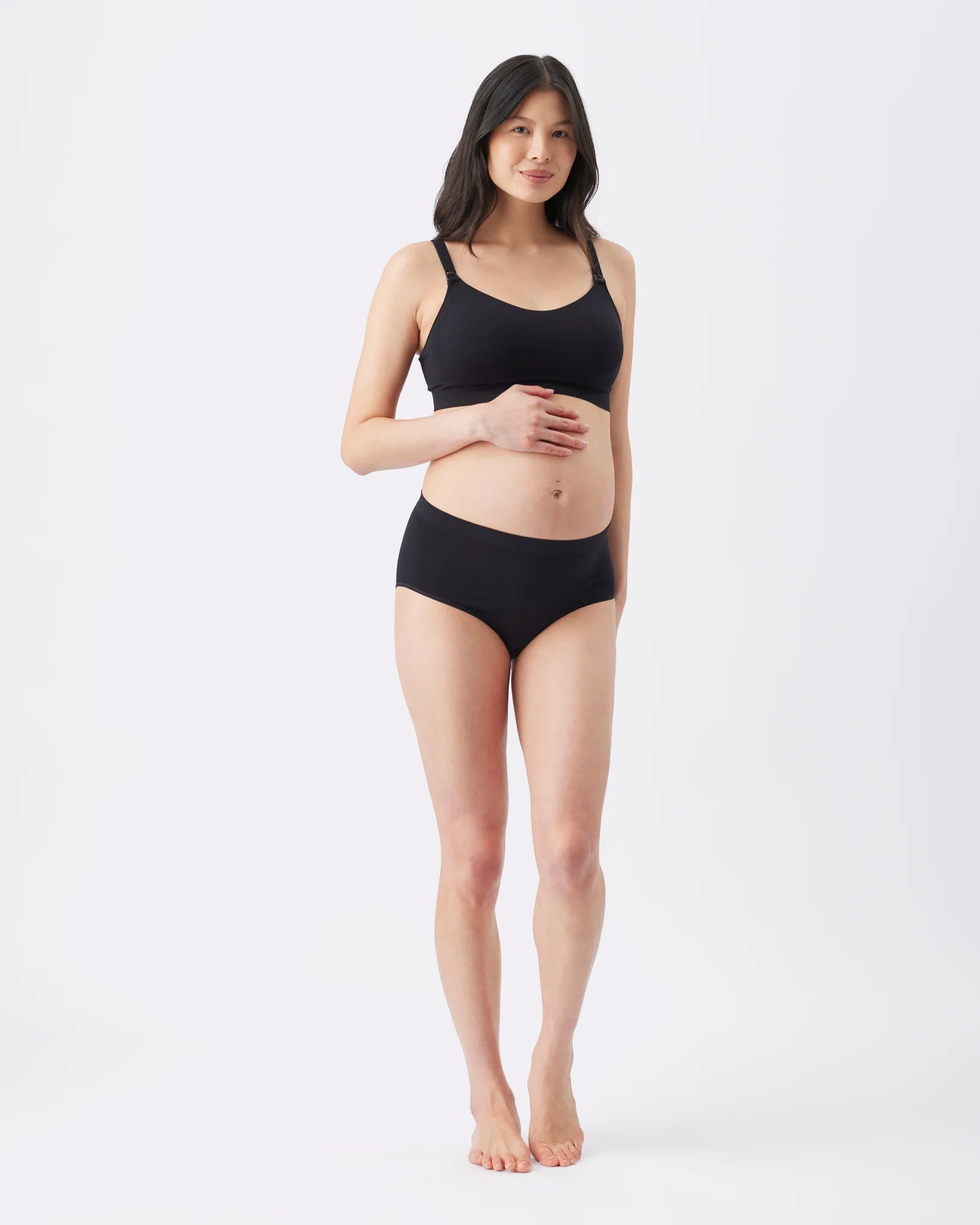 A pregnant woman modeling a black maternity bra and matching black underwear against a plain white studio background.