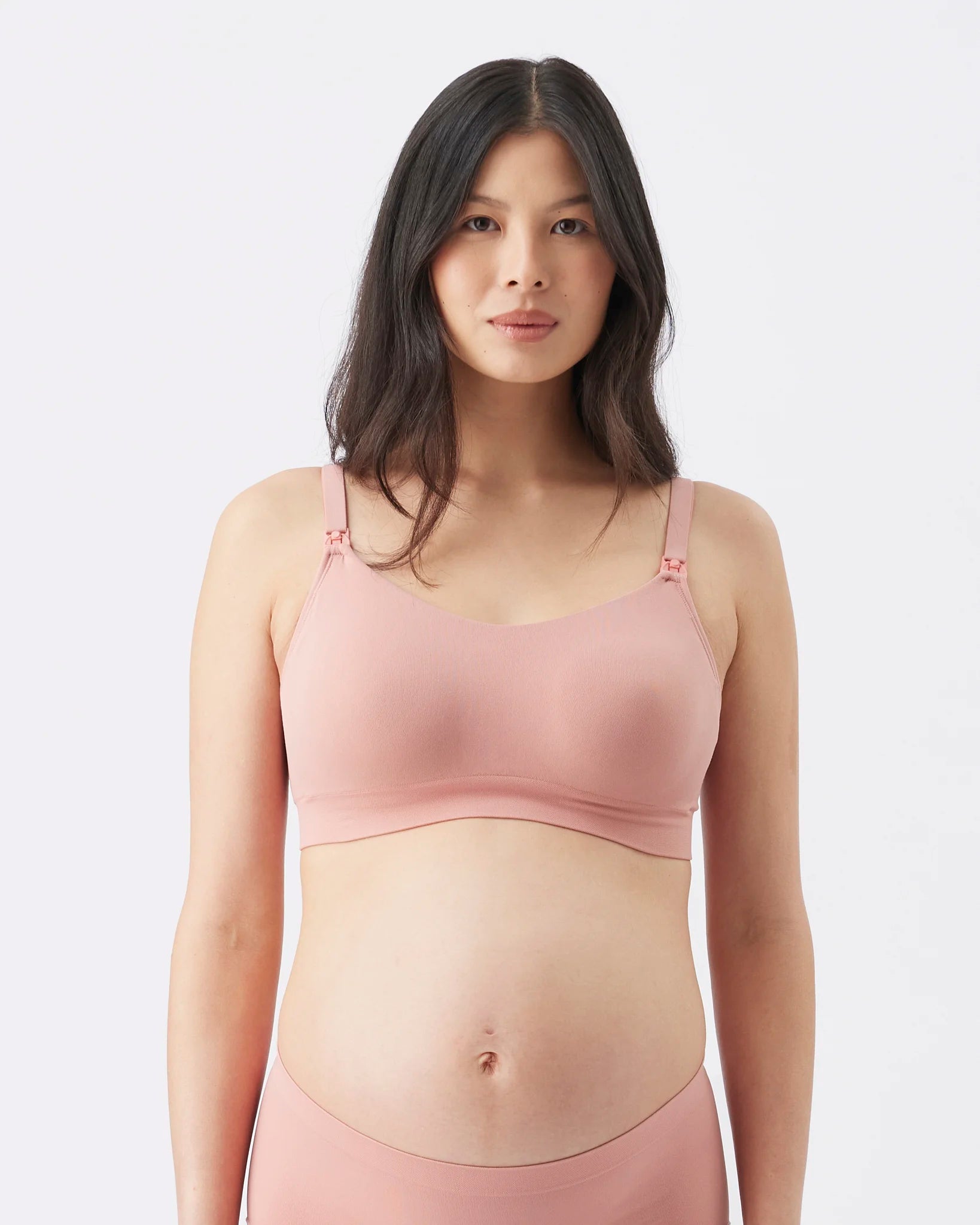 A pregnant model wearing a dusty pink maternity bra and matching high-waisted underwear against a light gray studio background.