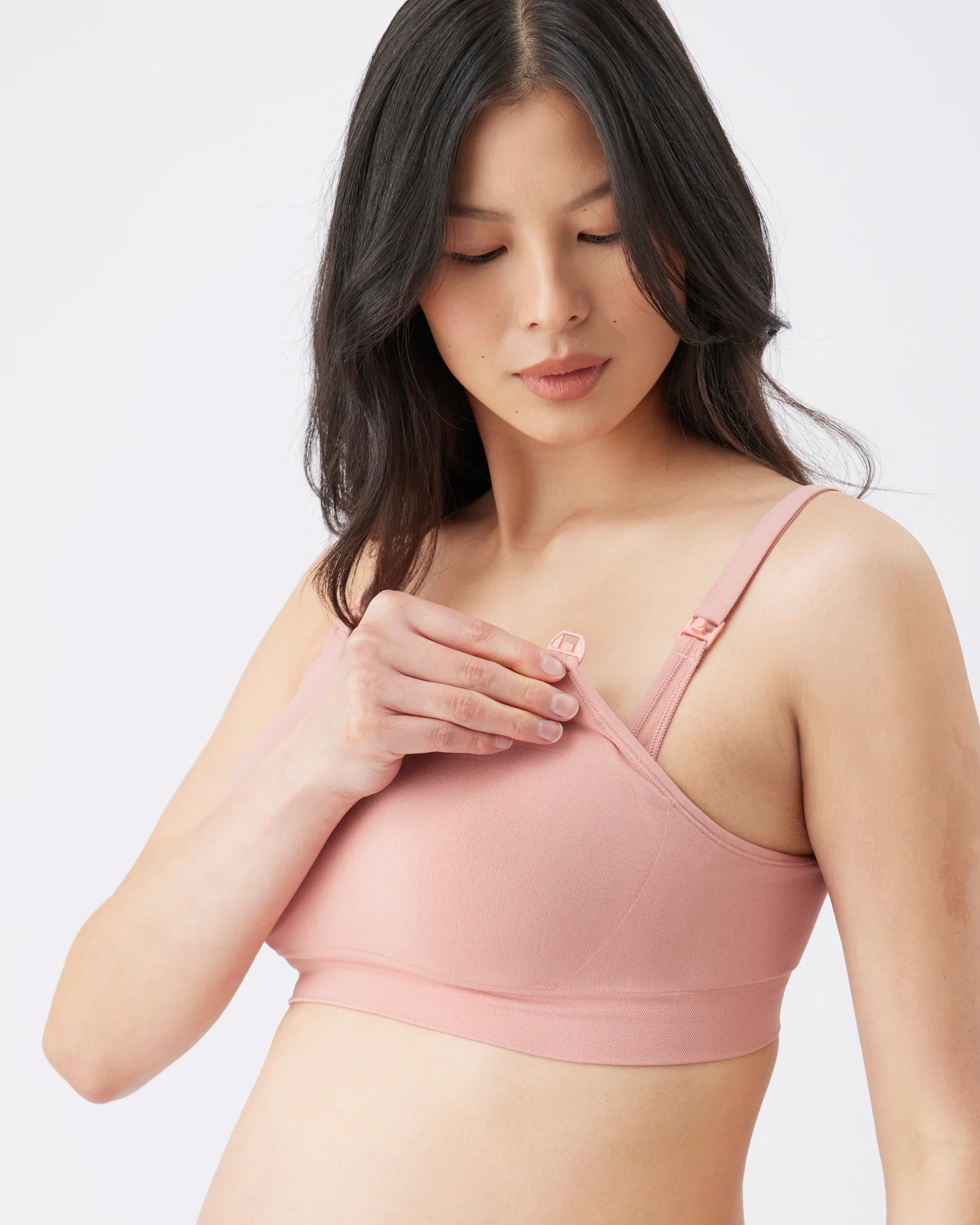 A woman wearing a dusty pink wireless nursing bra, adjusting the strap with her hand against a clean, light gray background.