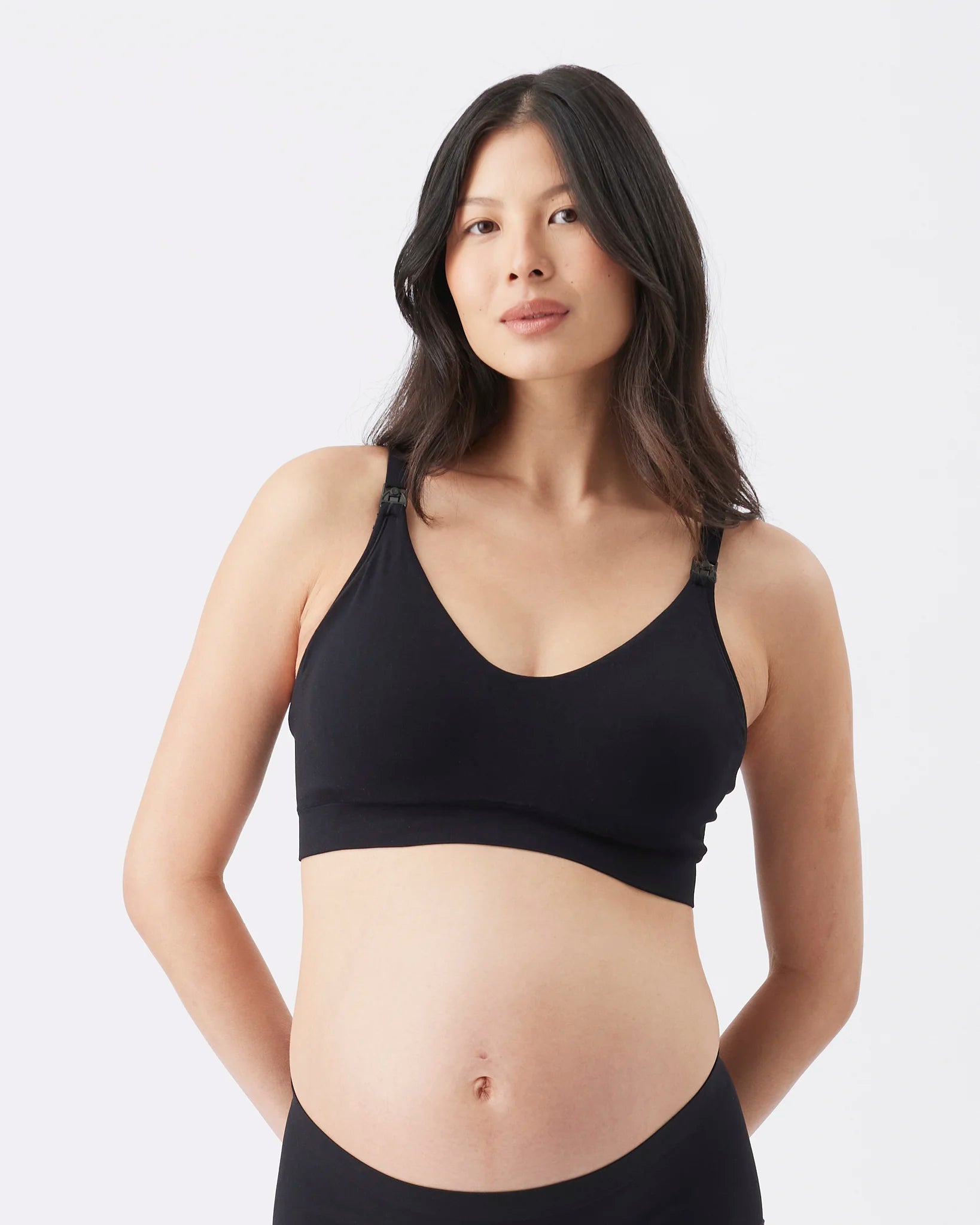A pregnant woman wearing a black maternity nursing bra and matching black underwear, photographed against a clean light gray studio background.