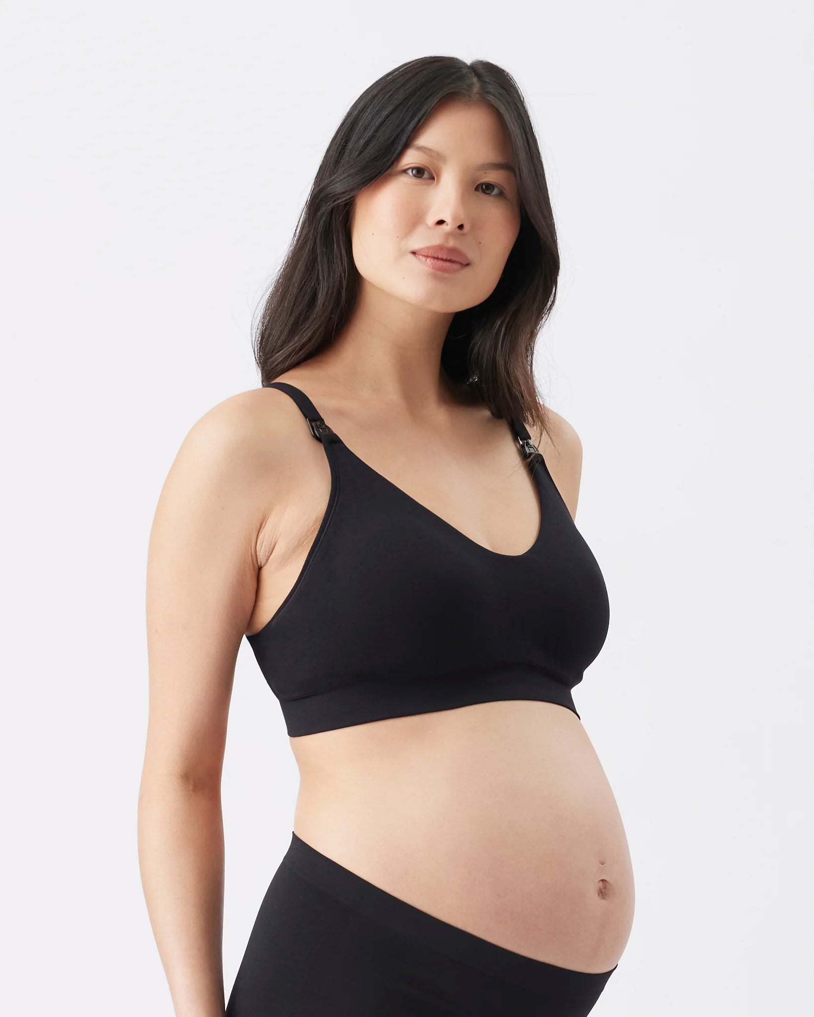A pregnant woman wearing a black maternity nursing bra and matching black underwear, photographed against a clean light gray studio background.