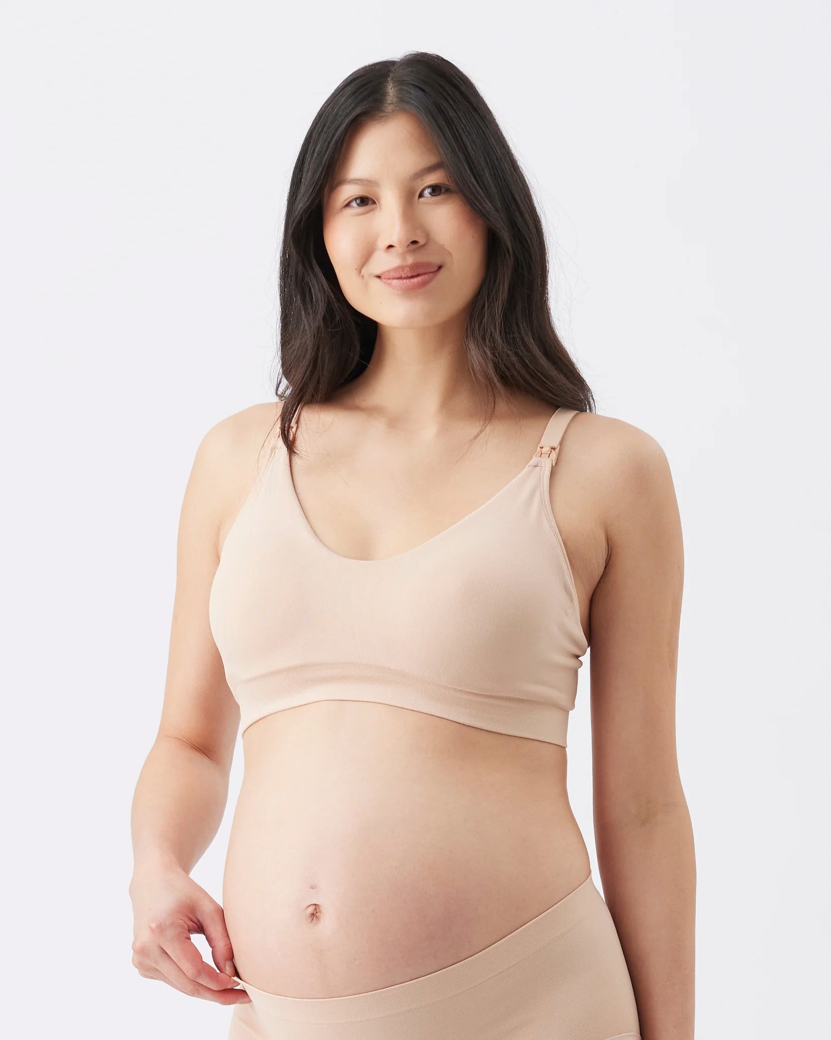A pregnant woman wearing a light beige maternity nursing bra and matching underwear, photographed against a clean white studio background.