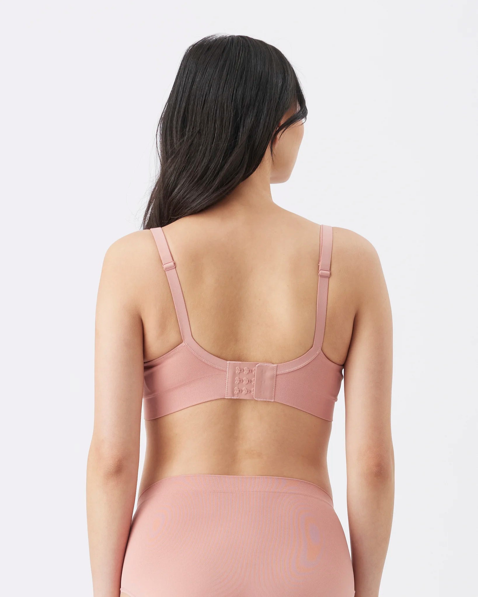 Rear view of a woman wearing a dusty pink seamless bra and matching high-waisted underwear against a light gray studio background.