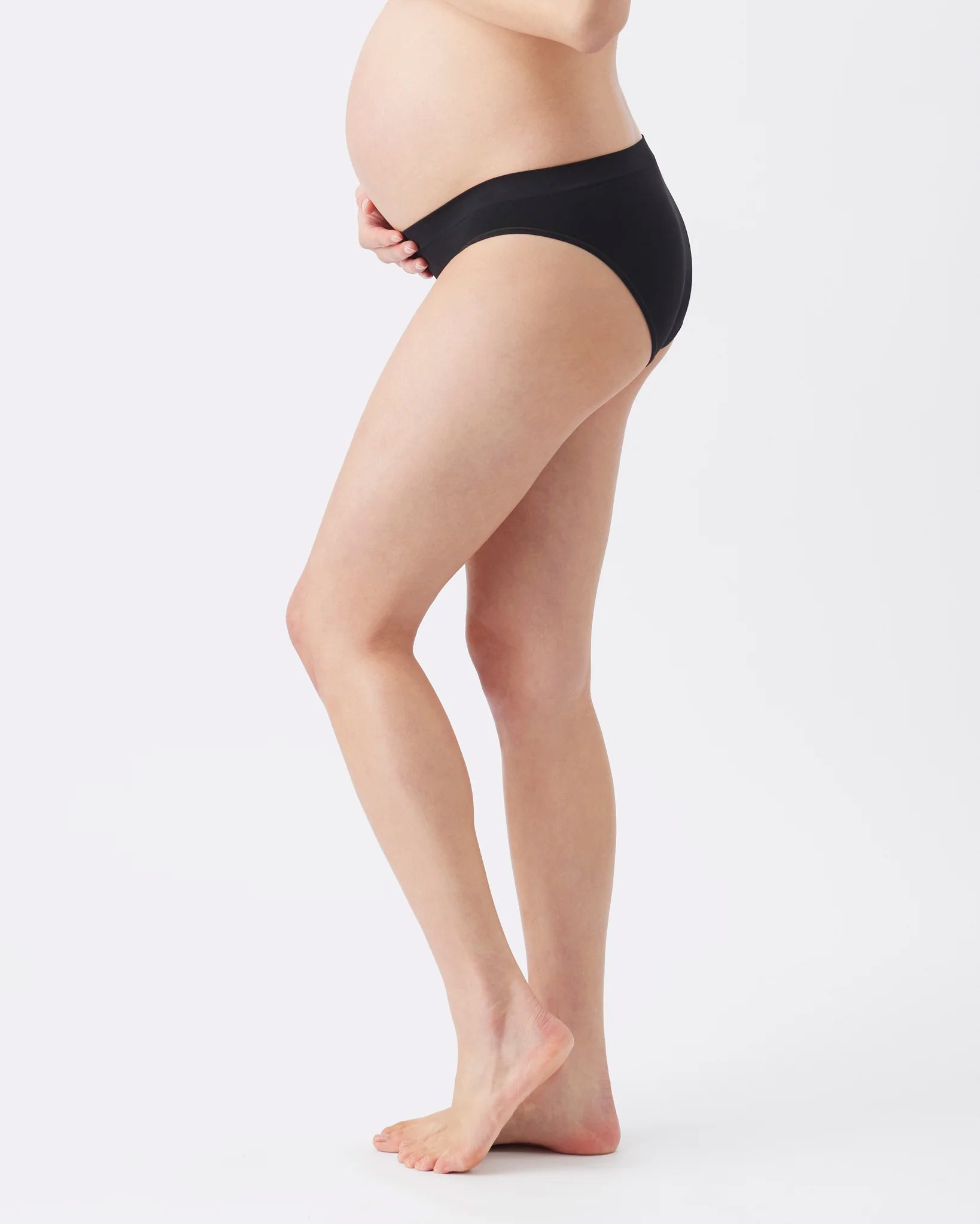 A pregnant model shown from the side wearing black maternity underwear, standing against a plain light gray studio background.