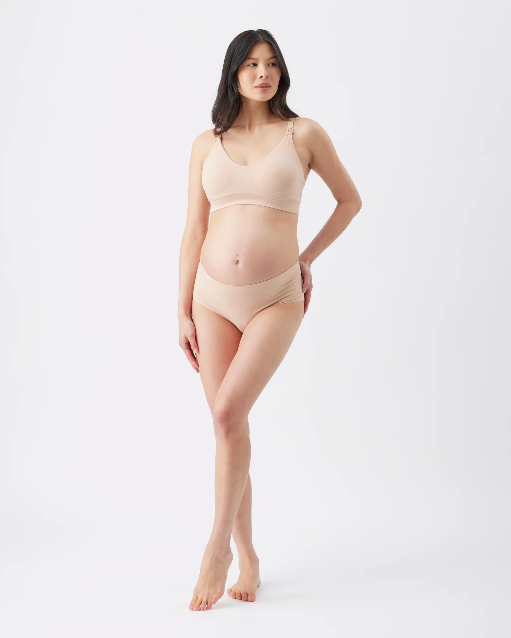A pregnant woman modeling a light beige maternity bra and matching underwear set against a plain white studio background.