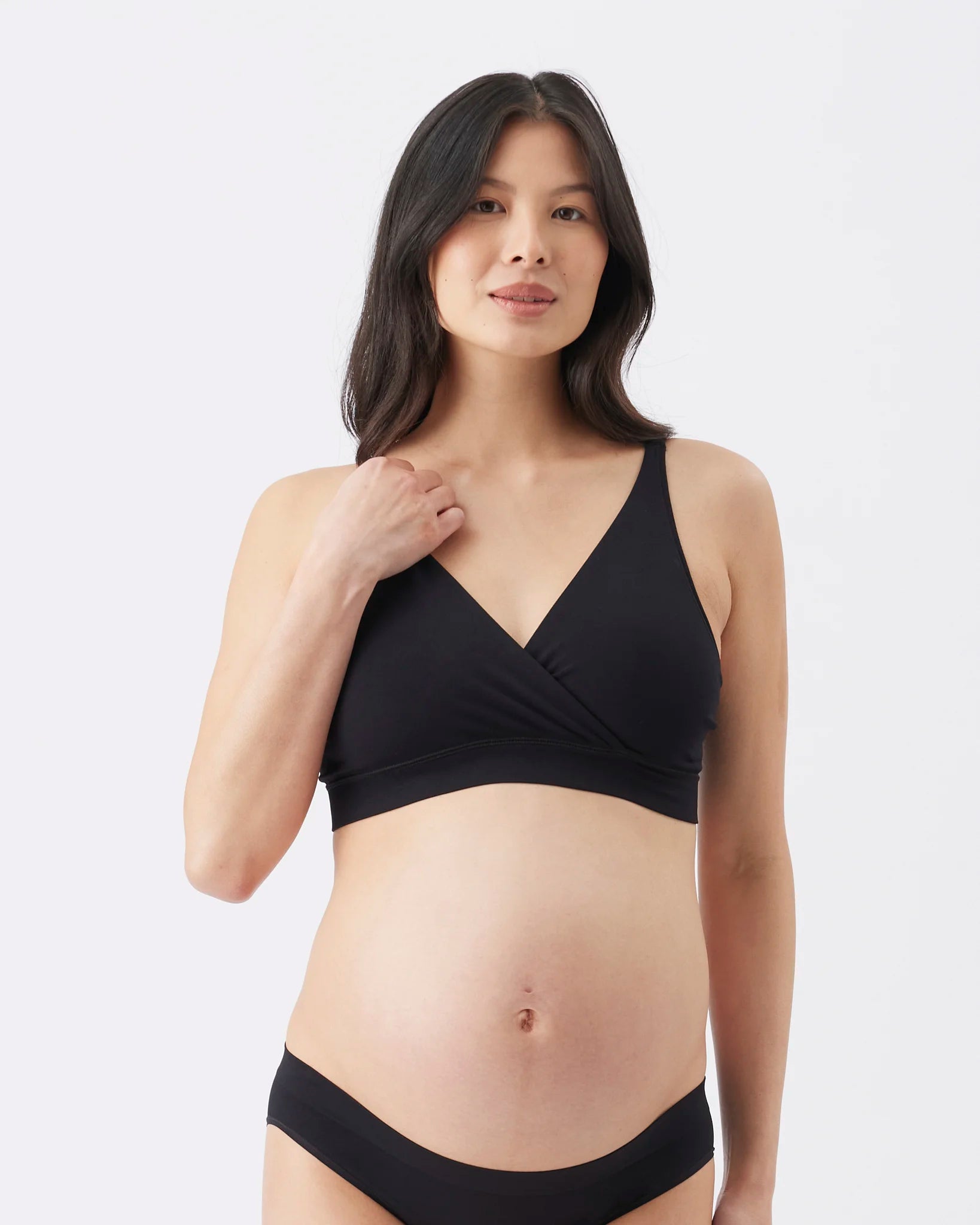 A pregnant model wearing a black maternity nursing bra and matching black underwear, standing against a light gray studio background.