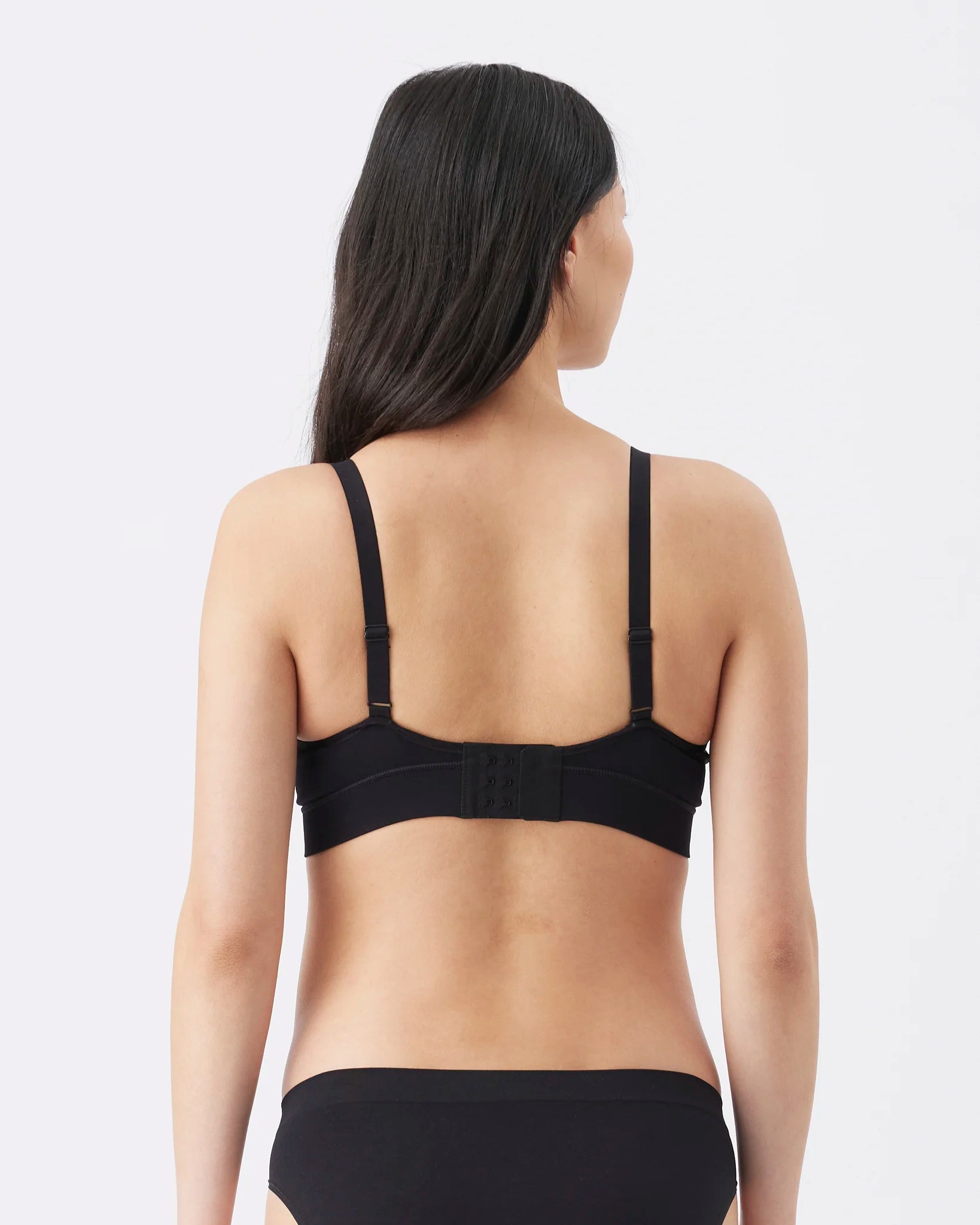 A woman shown from the back wearing a black bralette and black underwear against a light gray studio background.