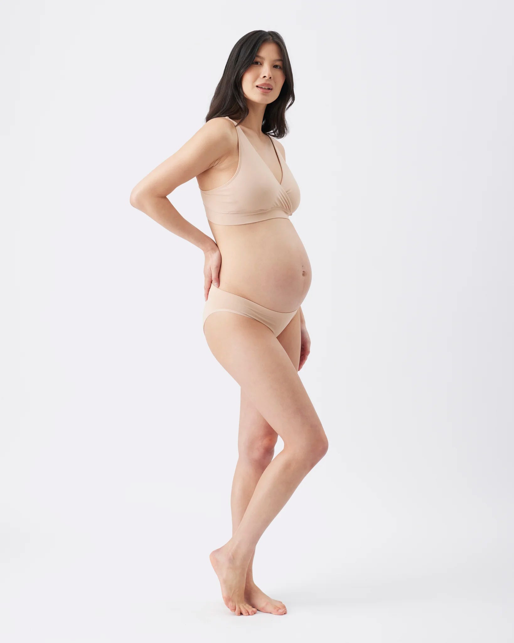 A pregnant woman modeling a matching beige maternity bra and underwear set against a plain white studio background.