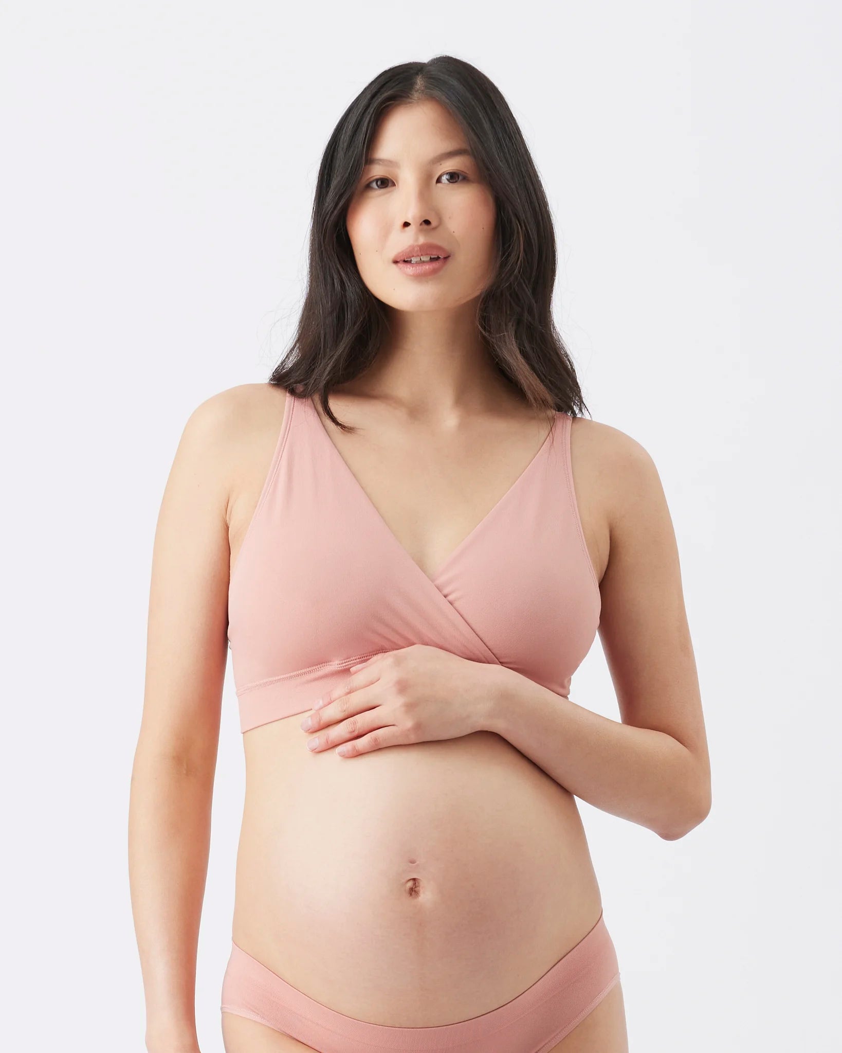 A pregnant woman wearing a dusty pink maternity bra and matching underwear, standing against a plain light gray studio background.