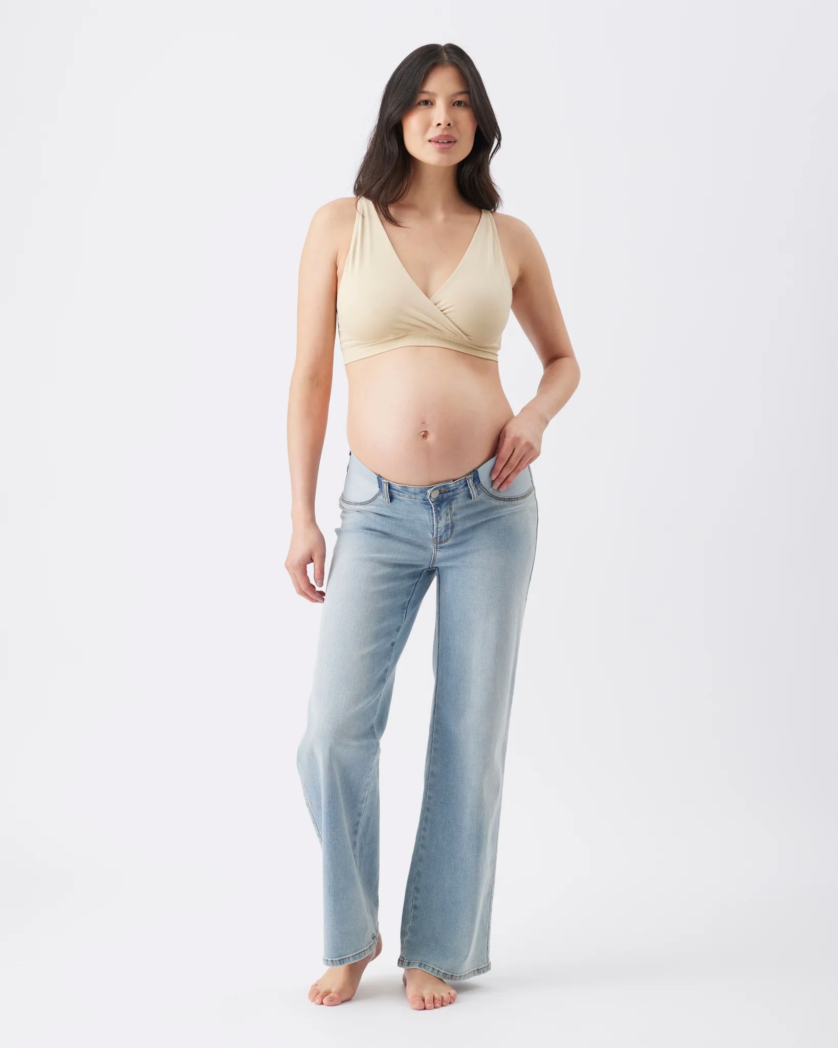 A pregnant woman stands barefoot against a plain white studio background, wearing a light beige wrap-style bikini top and light blue straight-leg jeans.