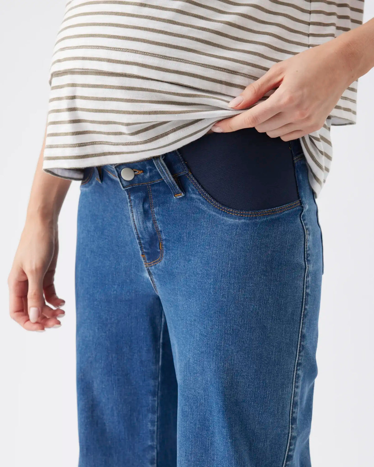Close-up of a person wearing blue jeans with a dark navy maternity belly band visible inside the front pocket, paired with a cream and gray striped top against a light gray background.