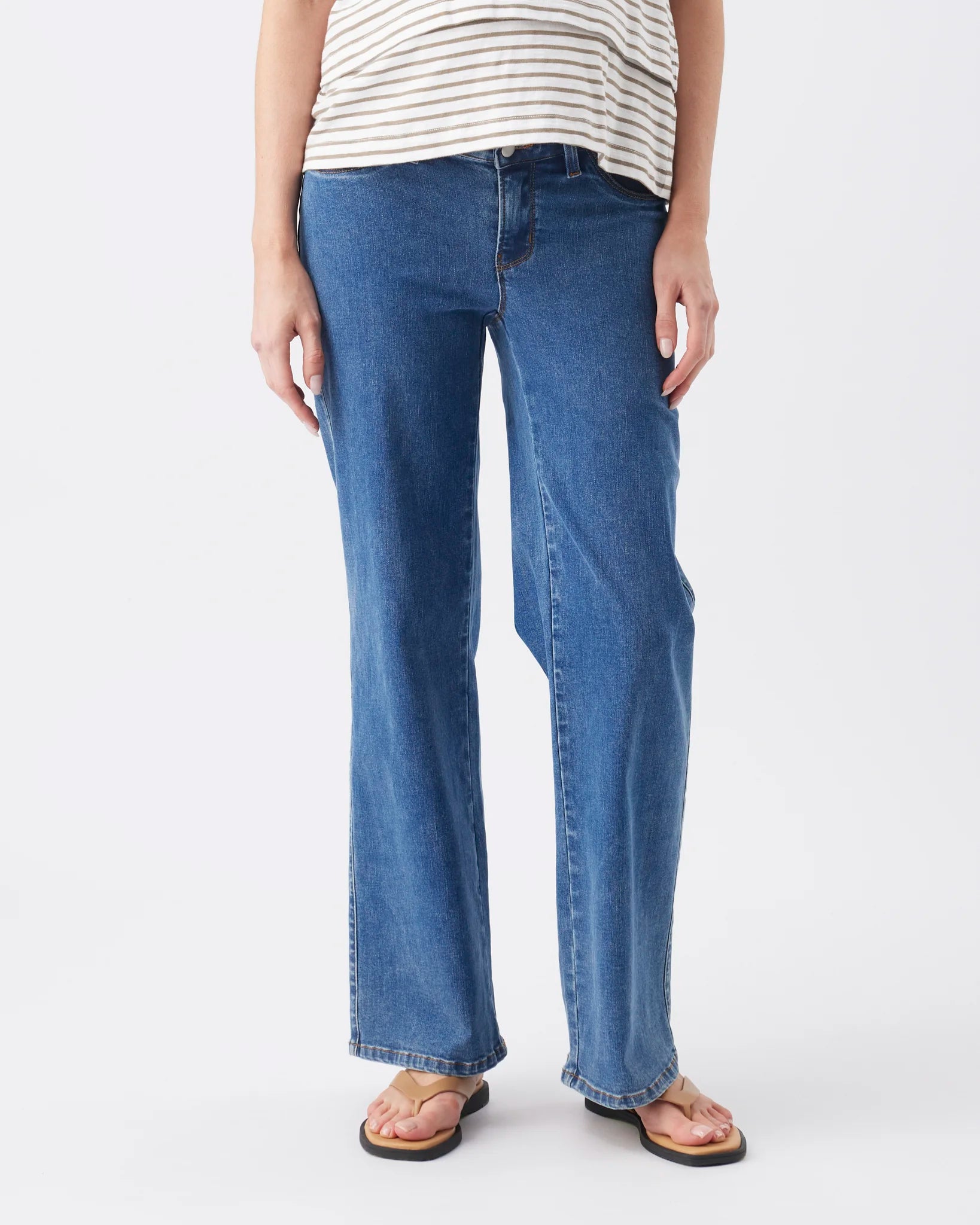 A person wearing medium-wash blue straight-leg jeans, a cream and navy striped top, and brown sandals, standing against a light gray studio background.