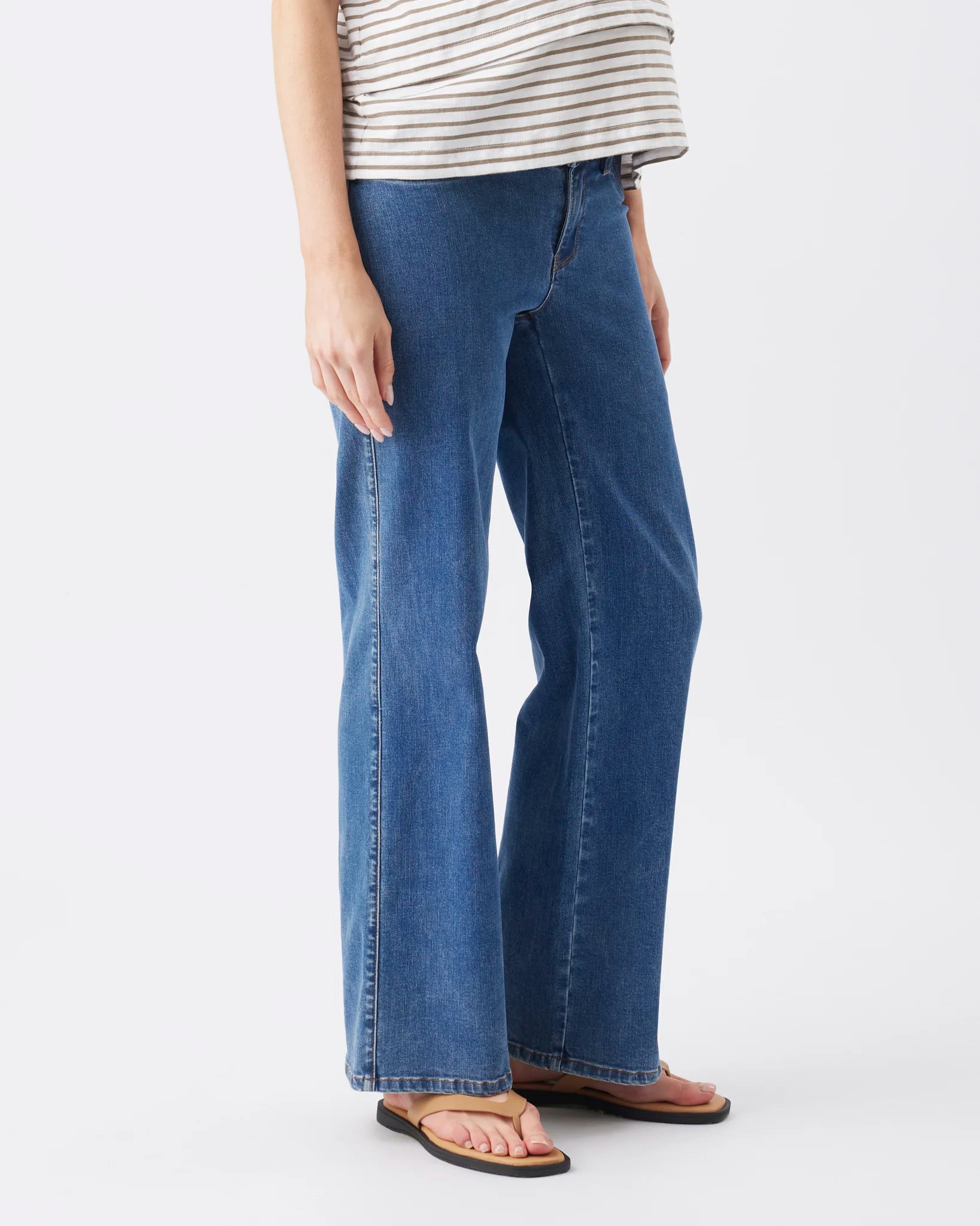 A person wearing medium-wash blue wide-leg jeans with a striped top and brown sandals, shown from the waist down against a light gray studio background.