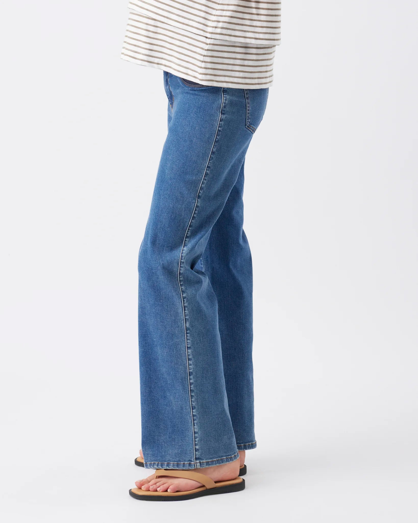A person shown from the waist down wearing medium-wash blue flare jeans, a cream and navy striped top, and tan sandals against a plain white studio background.