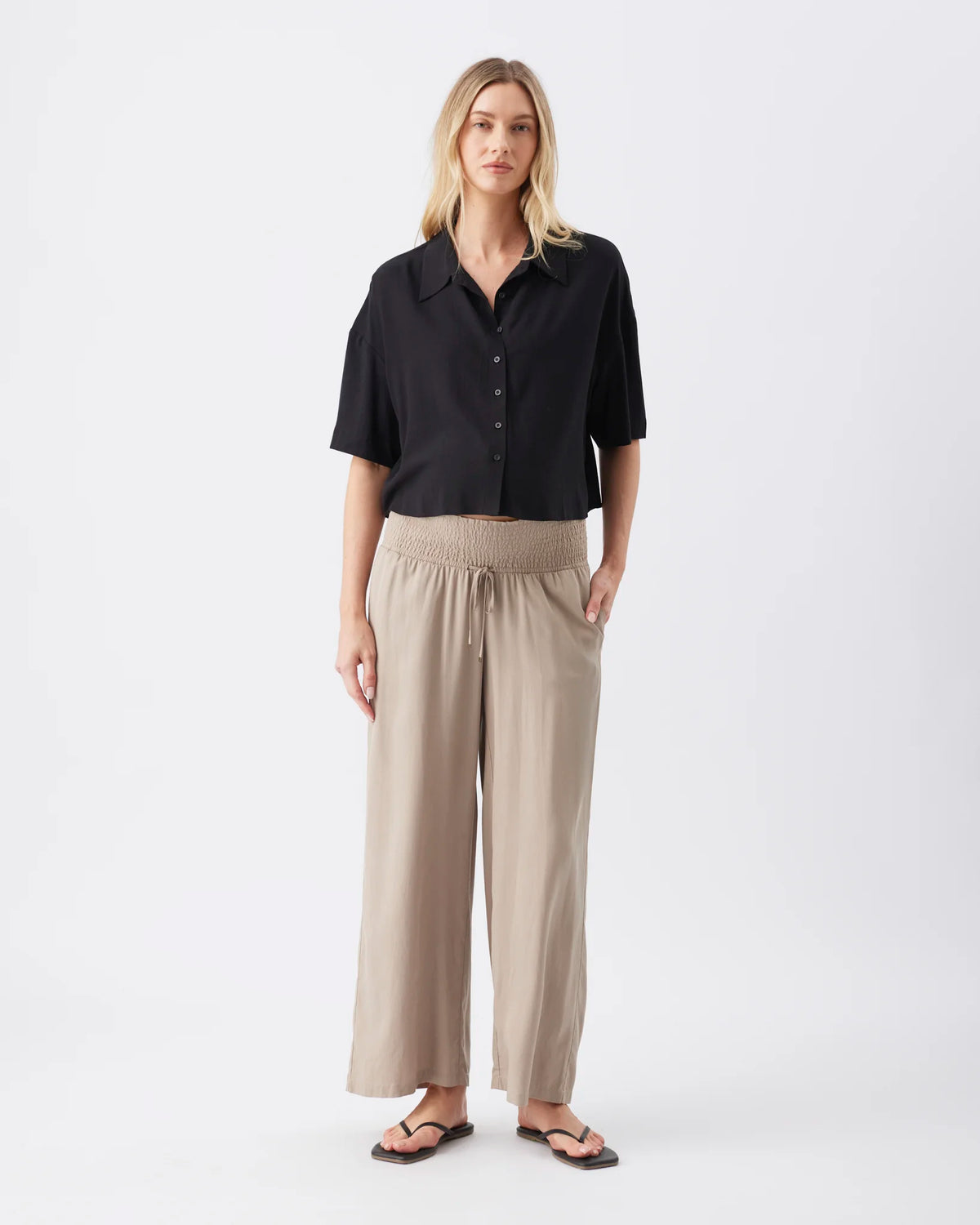 A woman modeling a black short-sleeve button-up shirt and beige wide-leg trousers against a plain light gray studio background.