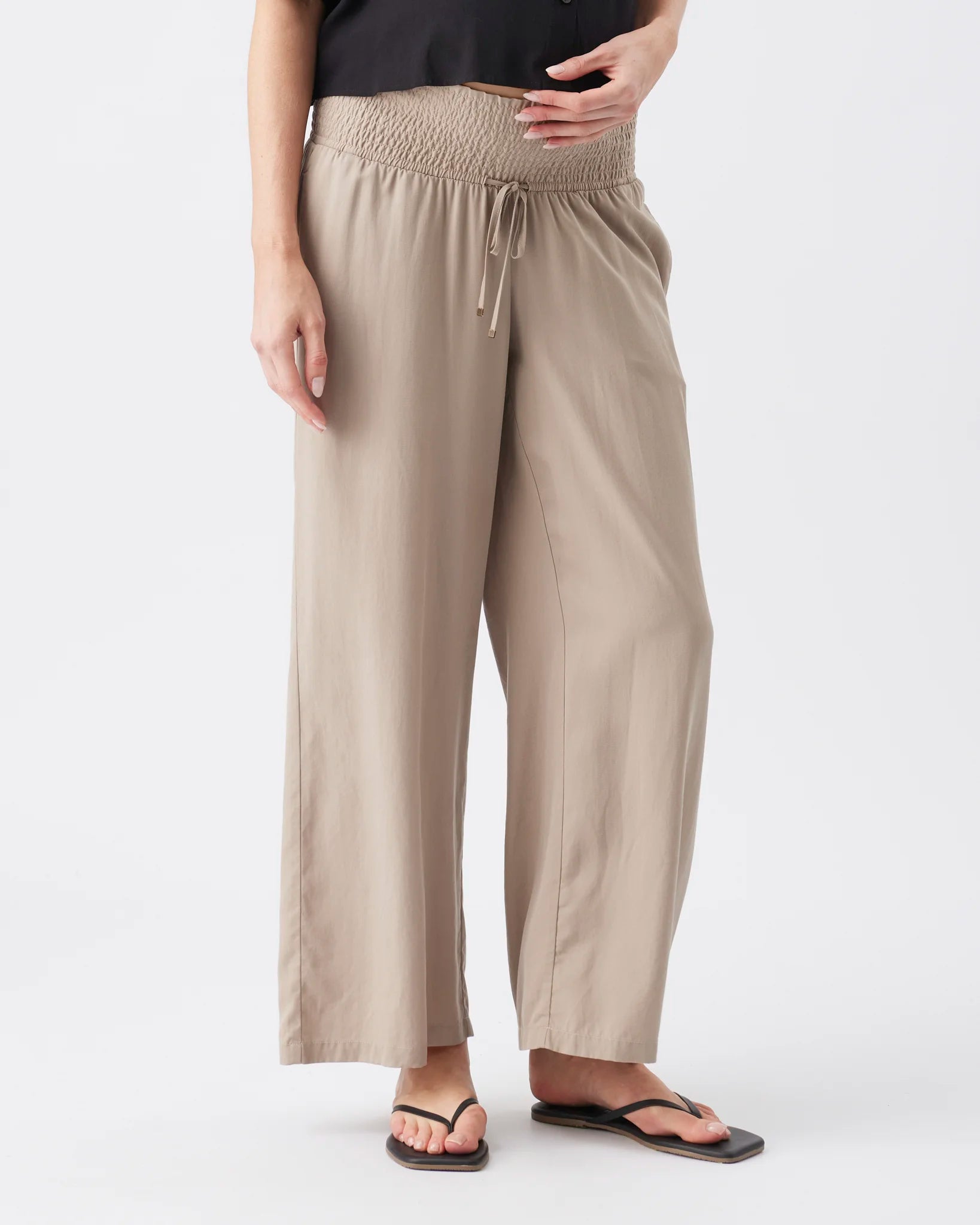 A person wearing loose, wide-leg beige pants with a smocked elastic waistband and drawstring, paired with a black top and black flip-flop sandals, against a plain light gray studio background.