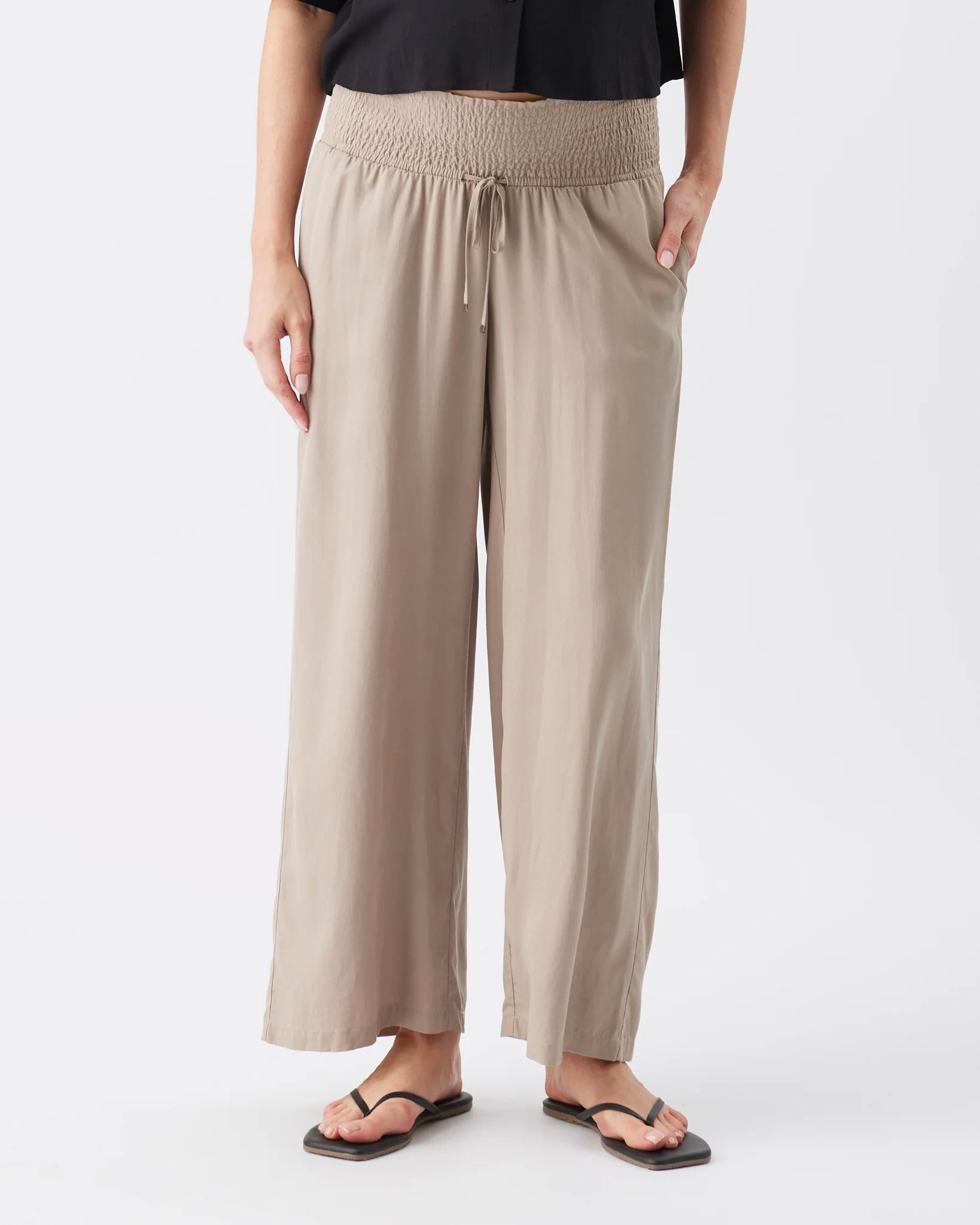 A person wearing loose, wide-leg cropped pants in a light taupe-beige color with an elastic waistband and drawstring, paired with black sandals against a plain light gray studio background.