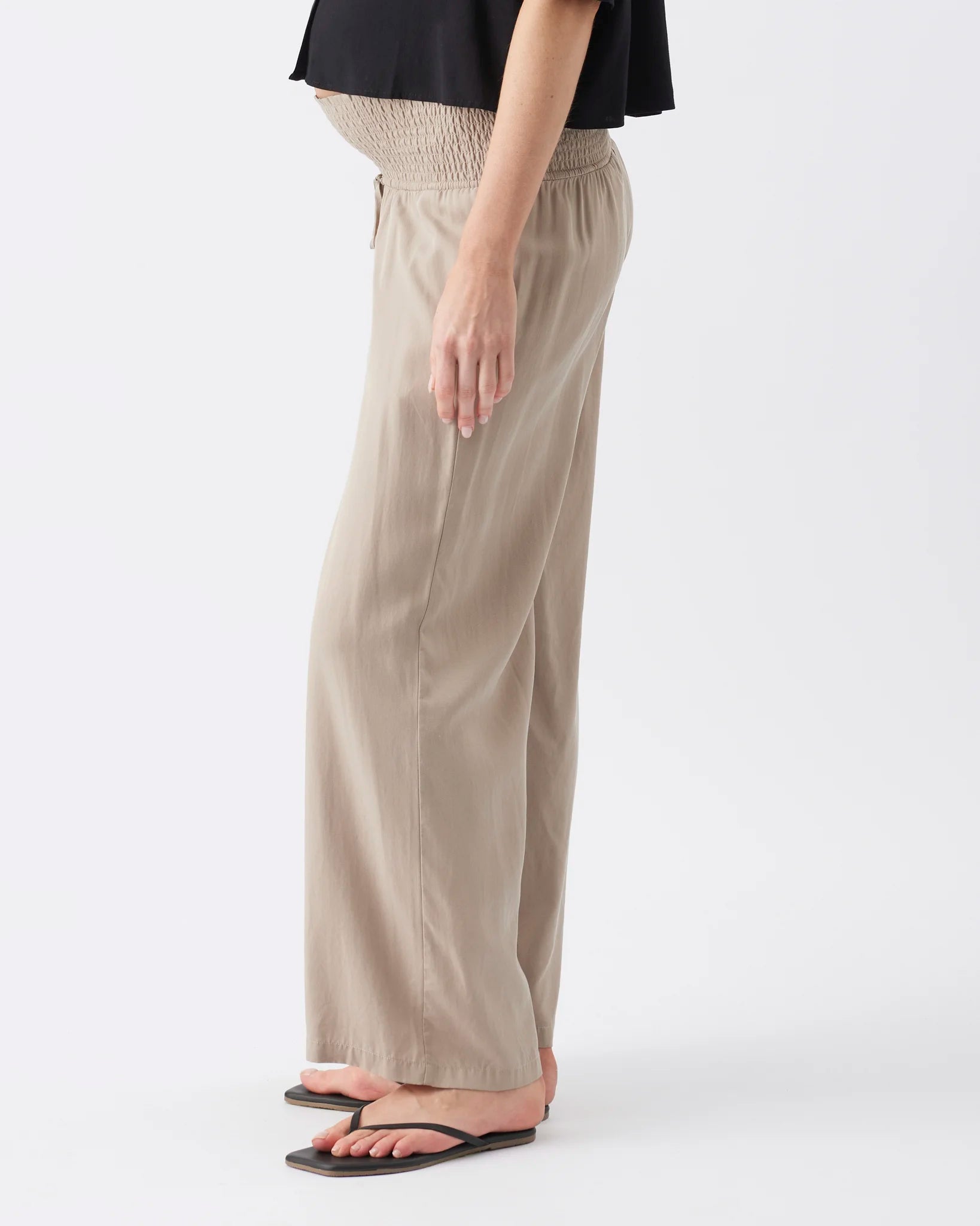 A side view of a pregnant model wearing beige wide-leg maternity pants with a smocked elastic waistband, paired with a black top and black flip-flops, against a plain white studio background.