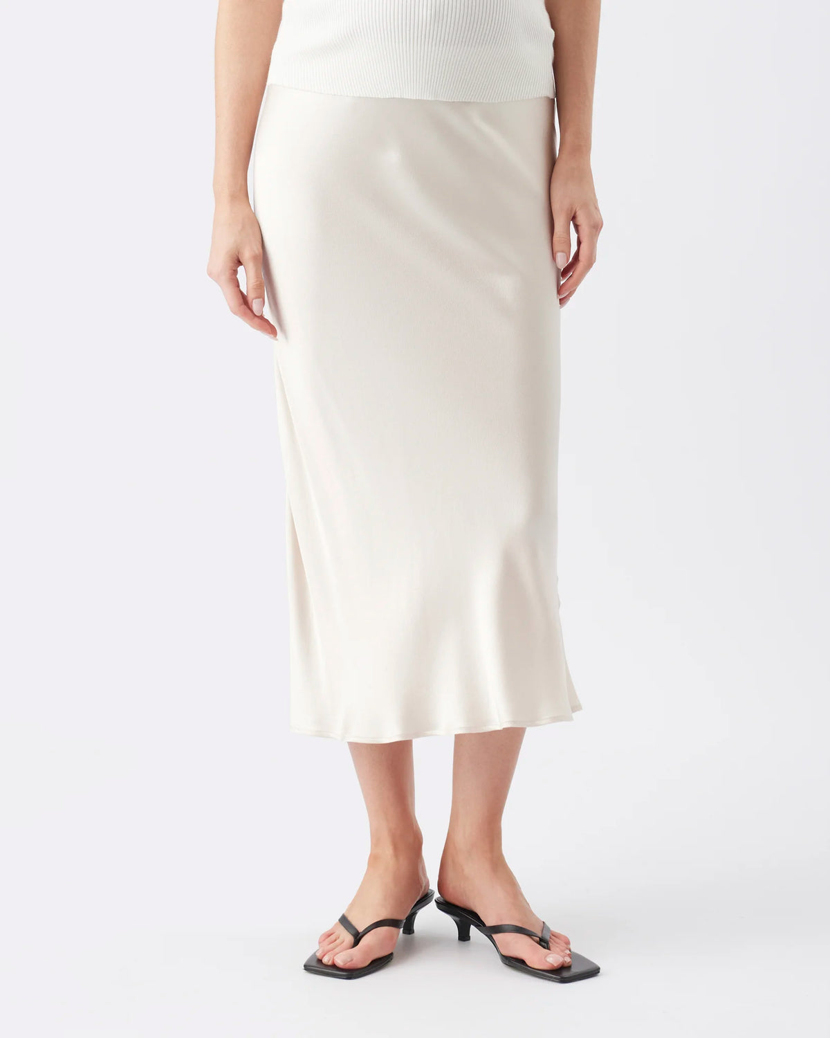 A model wearing a cream-colored satin midi skirt with a side slit, paired with black thong sandals and a white ribbed top, against a plain light gray background.