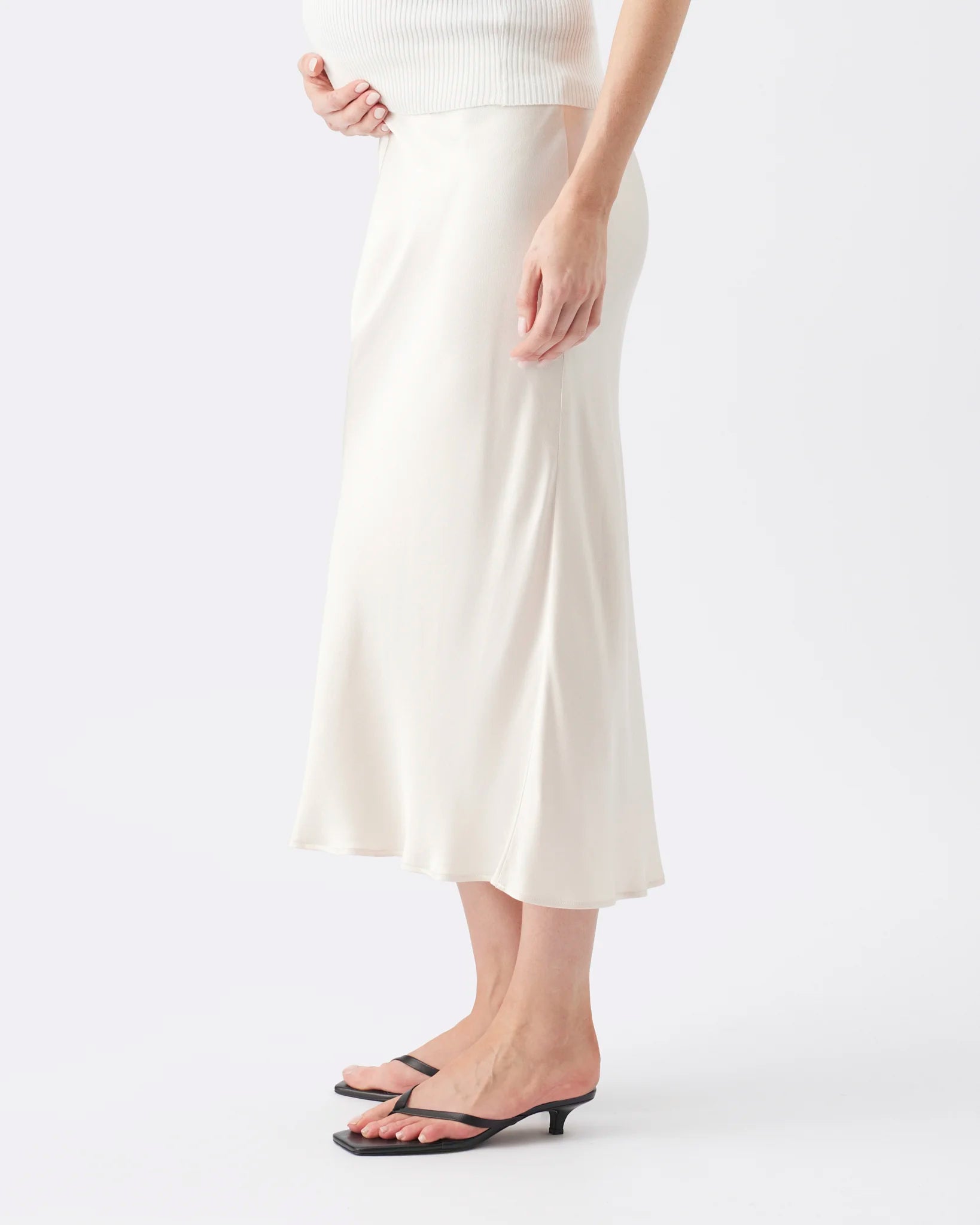 A model wearing a cream-colored satin midi skirt with a ribbed cream top and black flat sandals, shown from the waist down against a plain white studio background.