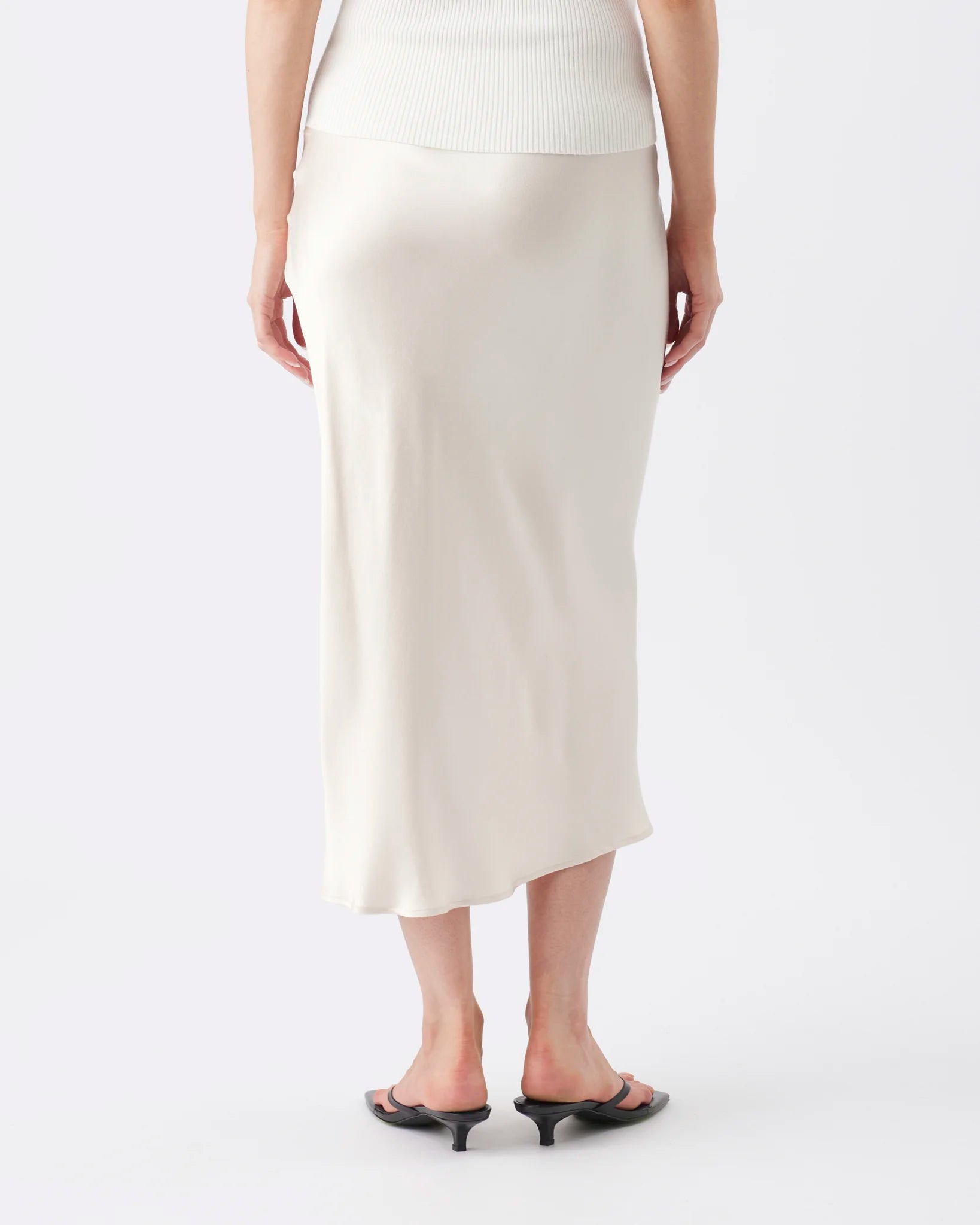 A model shown from the back wearing a cream-colored midi skirt, a ribbed white top, and black flat sandals against a plain white studio background.