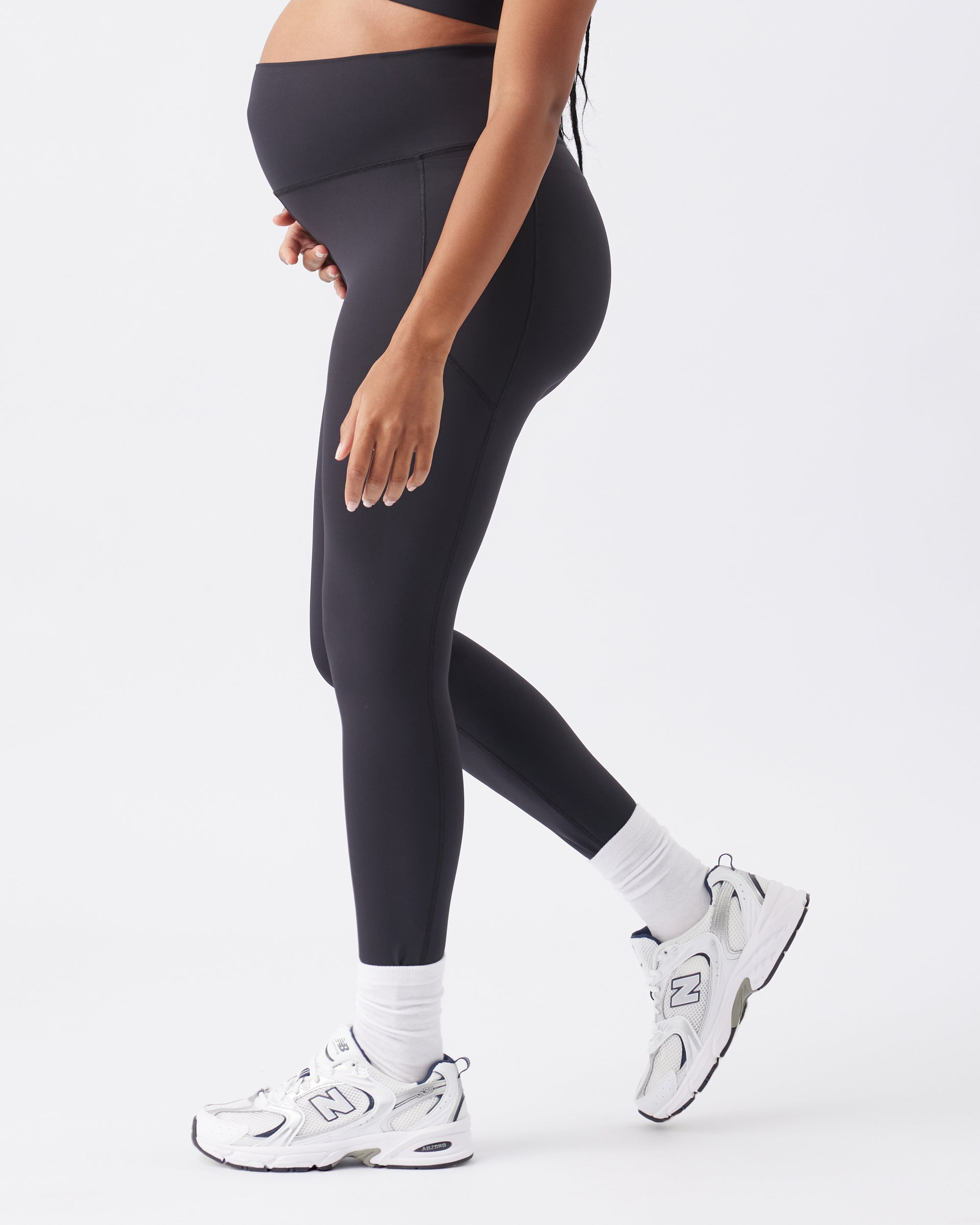 Side view of a pregnant woman wearing black leggings, white socks and white sneakers on a white background