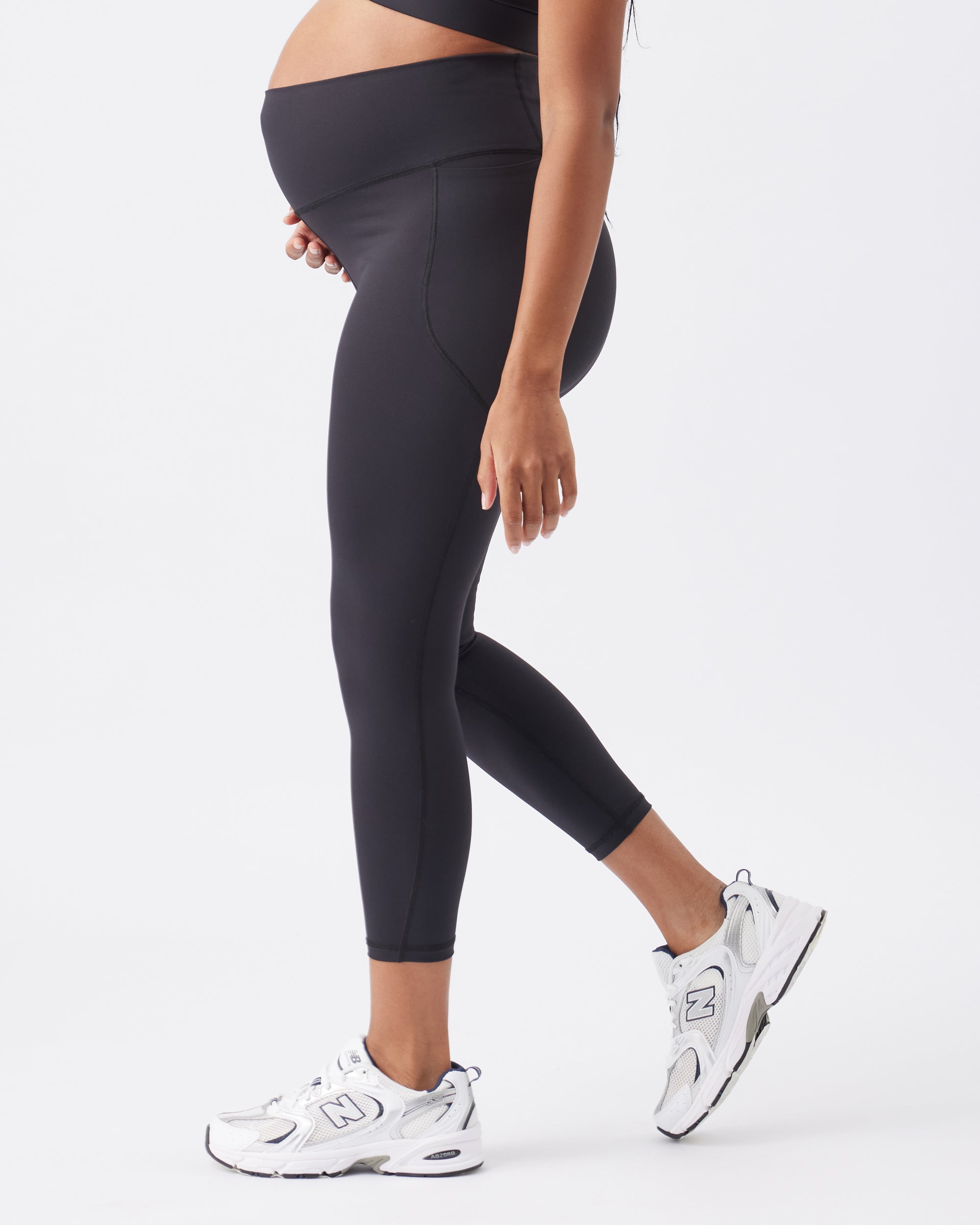 Side view of a pregnant woman wearing black leggings and white sneakers on a white background