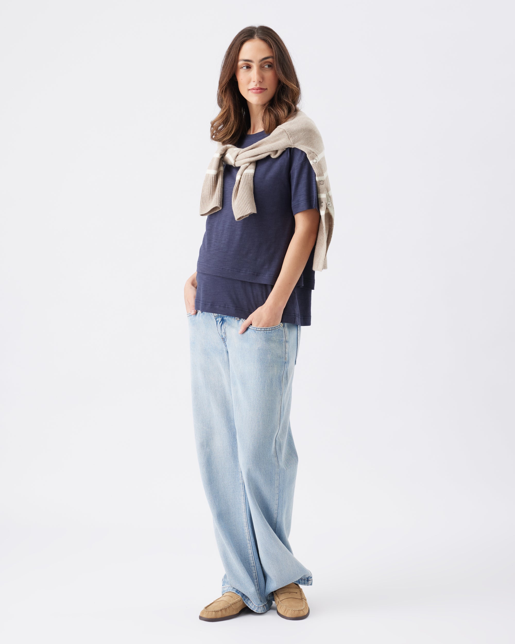 Woman wearing a navy top, light blue jeans, and a beige jumper tie dover the shoulders on a white background