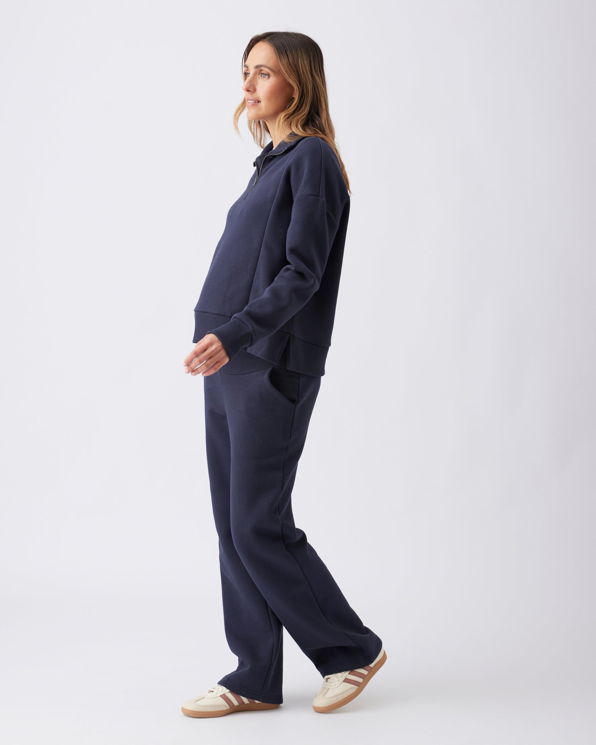 Full length side view of a pregnant woman wearing a navy blue tracksuit top and pants with sneakers.