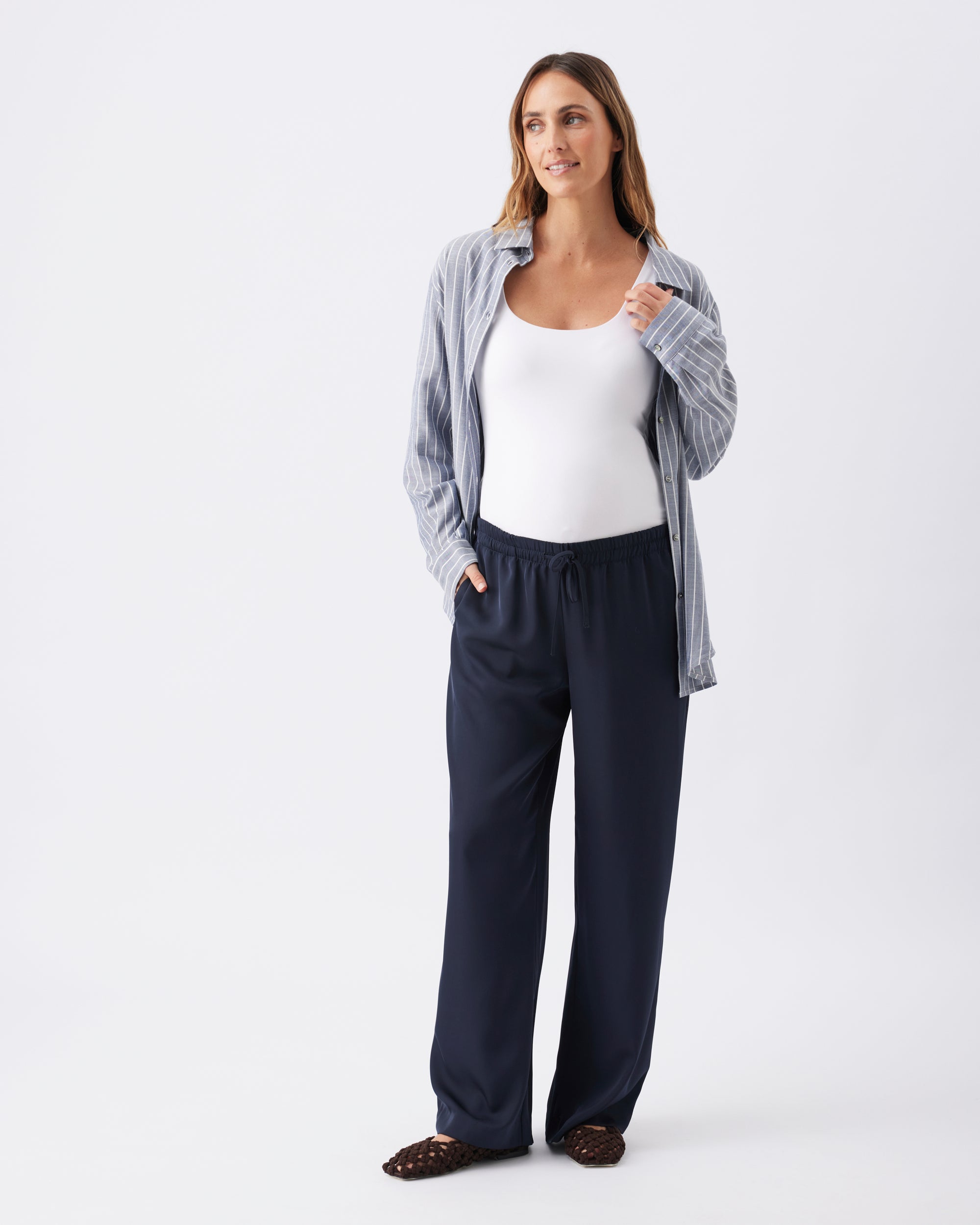 A pregnant woman wearing a white top, a light blue striped shirt unbuttoned over the top, and navy pants on a white background