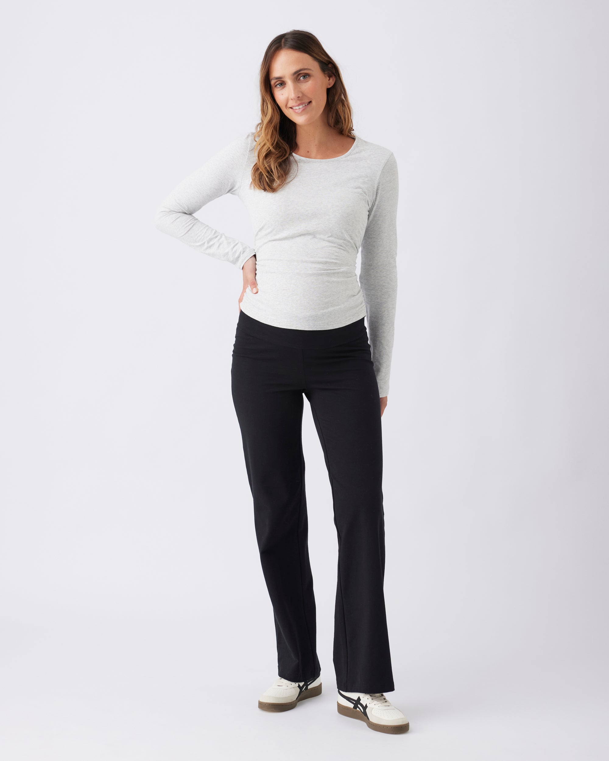 Full view woman wearing a light grey long-sleeve top and black pants on a white background